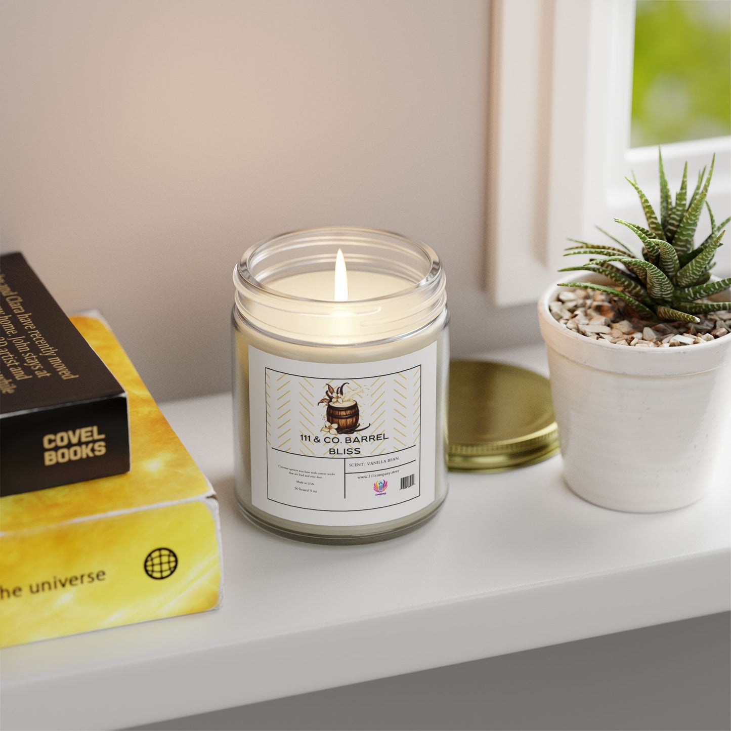 A coconut apricot wax candle labeled 111 & Company Barrel Bliss Vanilla Bean by 111 & Company rests in a clear 9oz jar on a white shelf with its golden lid nearby. It sits beside two books, one titled The Universe, and a potted succulent. Sunlight filters through the window, casting a soft glow.
