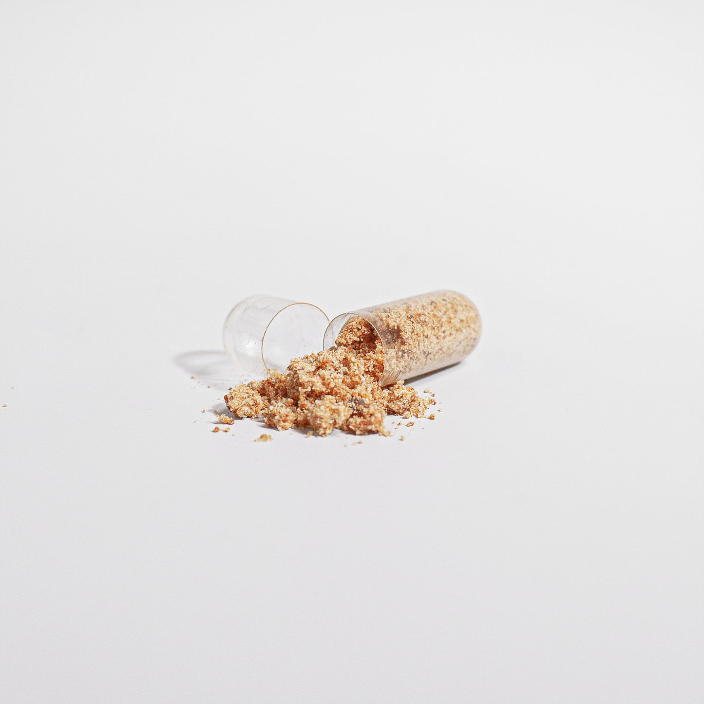 The 111 & COMPANY BEE PEARL capsule is open on a white surface, spilling its beige vitamin-rich powder. Finely textured granules slightly scatter around it, forming a small mound. Subtle shadows accentuate the minimalistic scene.