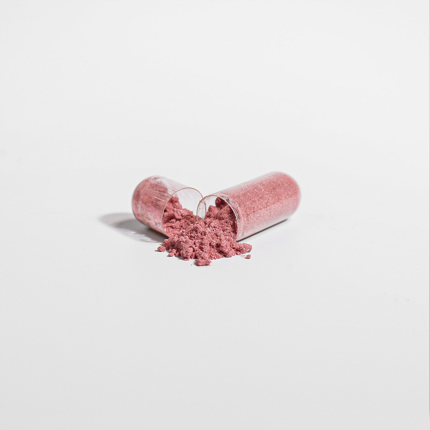 A pink powder spills from a split 111 & COMPANY BEETROOT CAPSULE on a white surface, revealing its vibrant contents. The beetroot-infused granules contrast with the smooth shell, showcasing their potential to enhance athletic performance through increased nitric oxide.