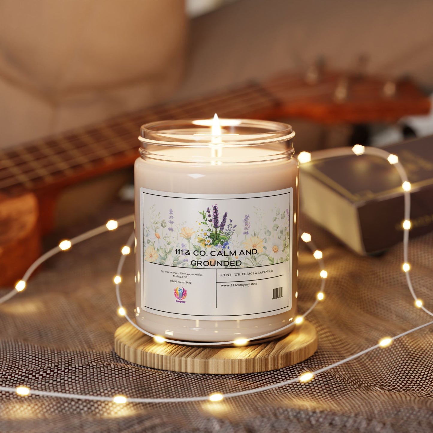 A lit 111 & Company Calm and Grounded 9oz soy candle with a White Sage & Lavender scent sits on a woven mat, surrounded by warm fairy lights. A ukulele and a closed book in the background enhance the cozy ambiance, featuring floral illustrations for relaxation and meditation.