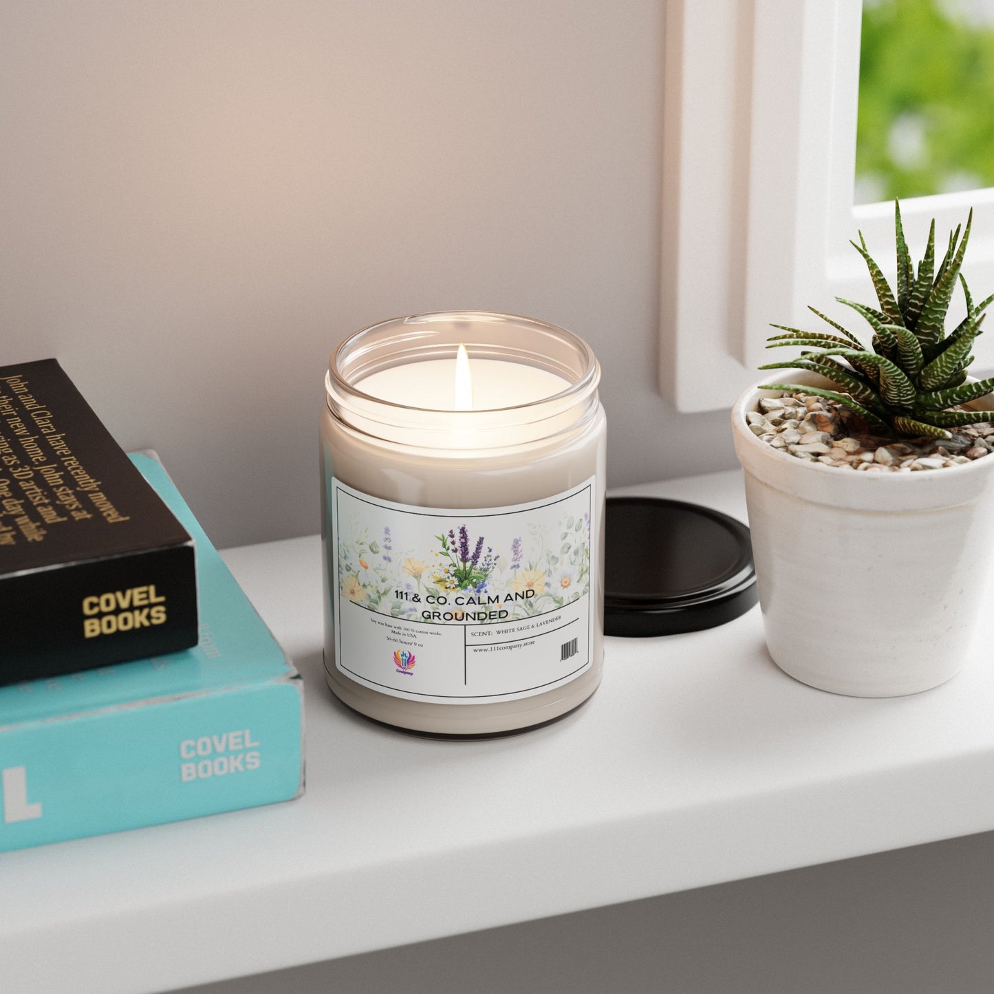 A 111 & Company Calm and Grounded White Sage & Lavender Soy Wax Candle by 111 & Company rests on a white shelf next to a small succulent. Its floral design and lavender scent promote relaxation, as books with visible titles bask in sunlight streaming through the window.