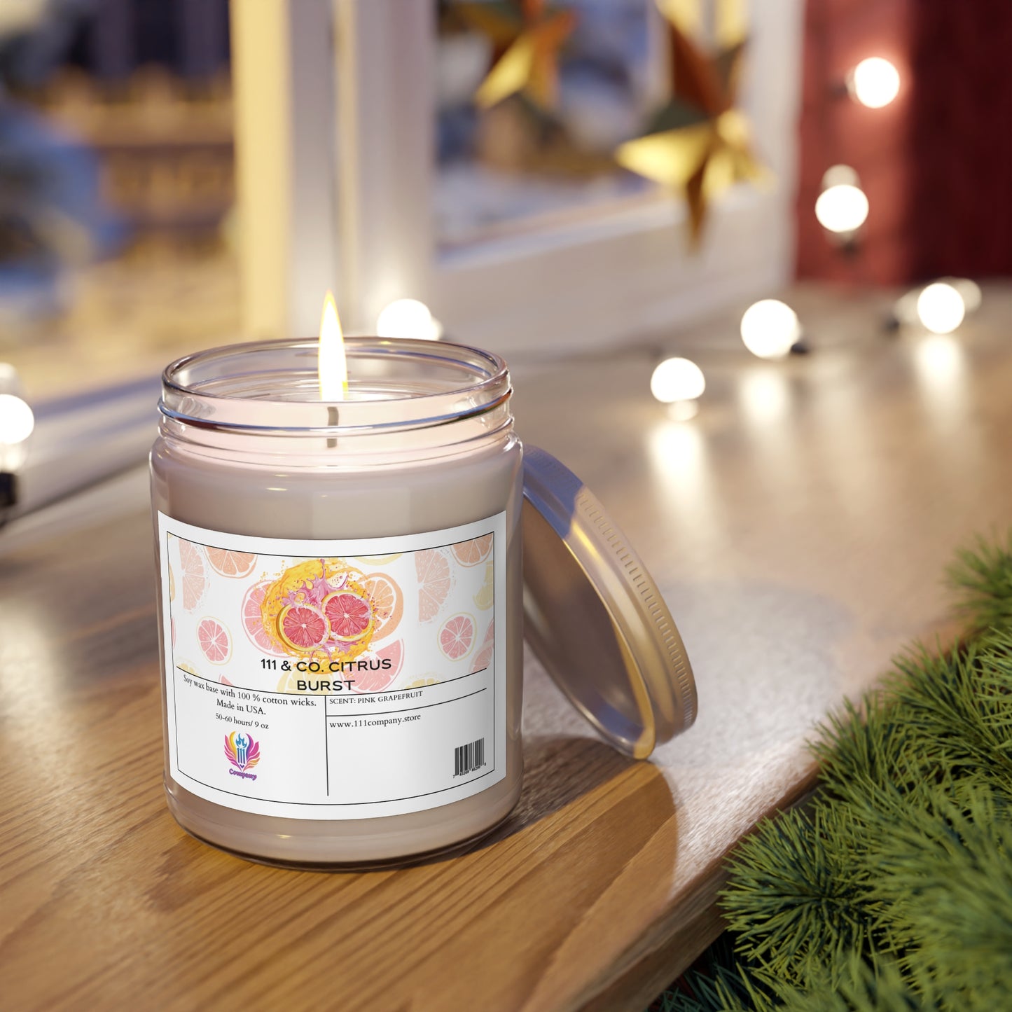 A 111 & COMPANY CITRUS BURST soy wax candle with a pink grapefruit scent from 111 & Company sits in a jar on wood. Its label shows citrus slices. A window features star decorations and blurred string lights, creating a cozy feel. Greenery is visible to the right.