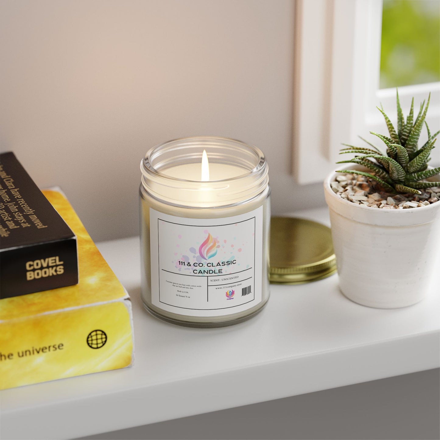 A lit 111 & Company Classic Unscented Candle in a clear jar sits on a white shelf. Made from eco-friendly coconut apricot wax, it emits a calming glow. Nearby, a potted succulent and two yellow books—one titled Covel Books, the other with planet imagery—enhance the decor as daylight filters through the window.