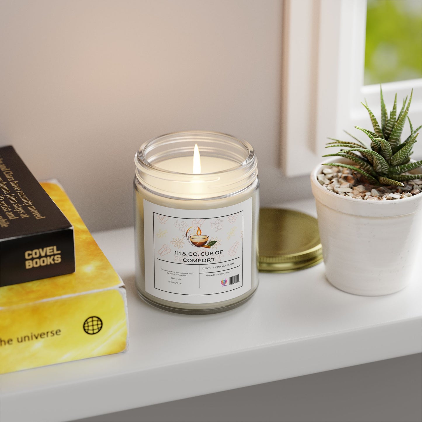 A lit 111 & Company Cup of Comfort Cinnamon Chai Candle in a clear jar with coconut apricot wax sits on a white windowsill by a potted succulent. Nearby, two stacked books with yellow and black covers. Outside, the view is bright and blurred. A golden lid lies beside this aromatic escape.