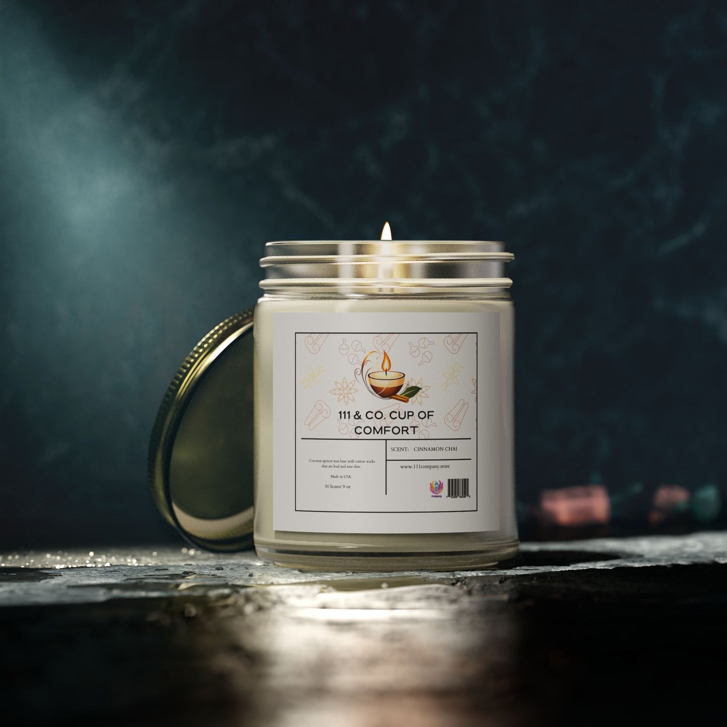 A lit 9oz 111 & Company Cup of Comfort cinnamon chai candle with a gold lid and cup illustration sits on a dark surface. Made with eco-friendly coconut apricot wax, it exudes an aromatic escape amid blurred hints of pink and blue objects in the dim light.