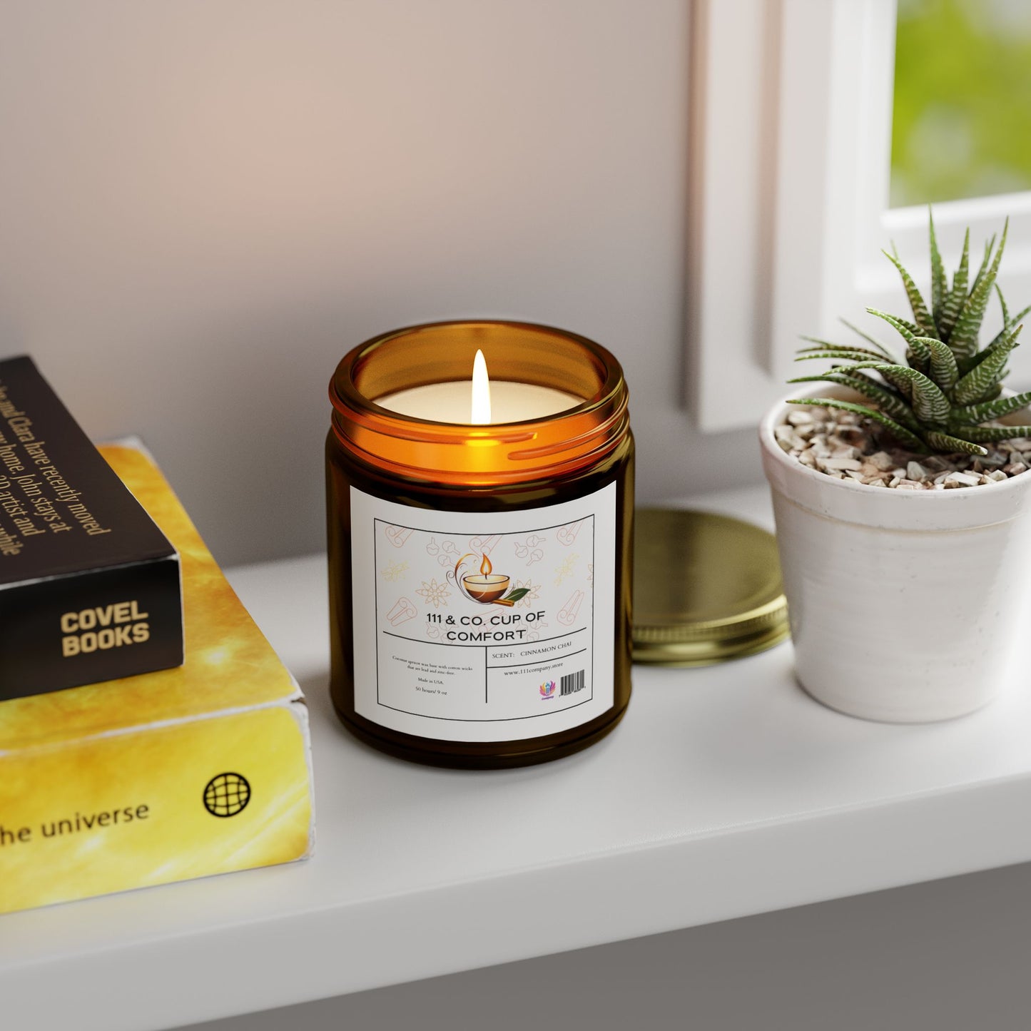 An amber jar labeled 111 & Company Cup of Comfort Cinnamon Chai Scented Candle by 111 & Company rests on a white surface. Nearby, two stacked books, including a yellow one titled The Universe, complement the scene with a small potted succulent near the window.