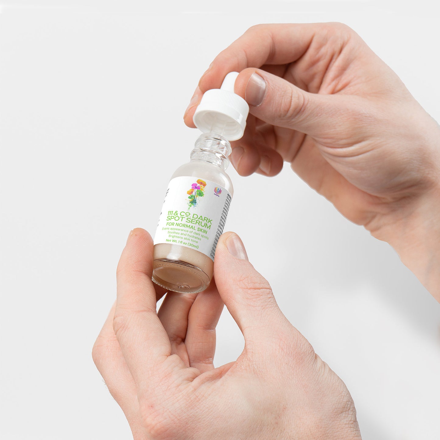 Two hands hold a small glass bottle with a white dropper cap. The label reads 111 & COMPANY DARK SPOT SERUM FOR NORMAL SKIN,” featuring colorful text and illustrations. The bottle has a light brown liquid promising dark spot reduction against a plain white background.