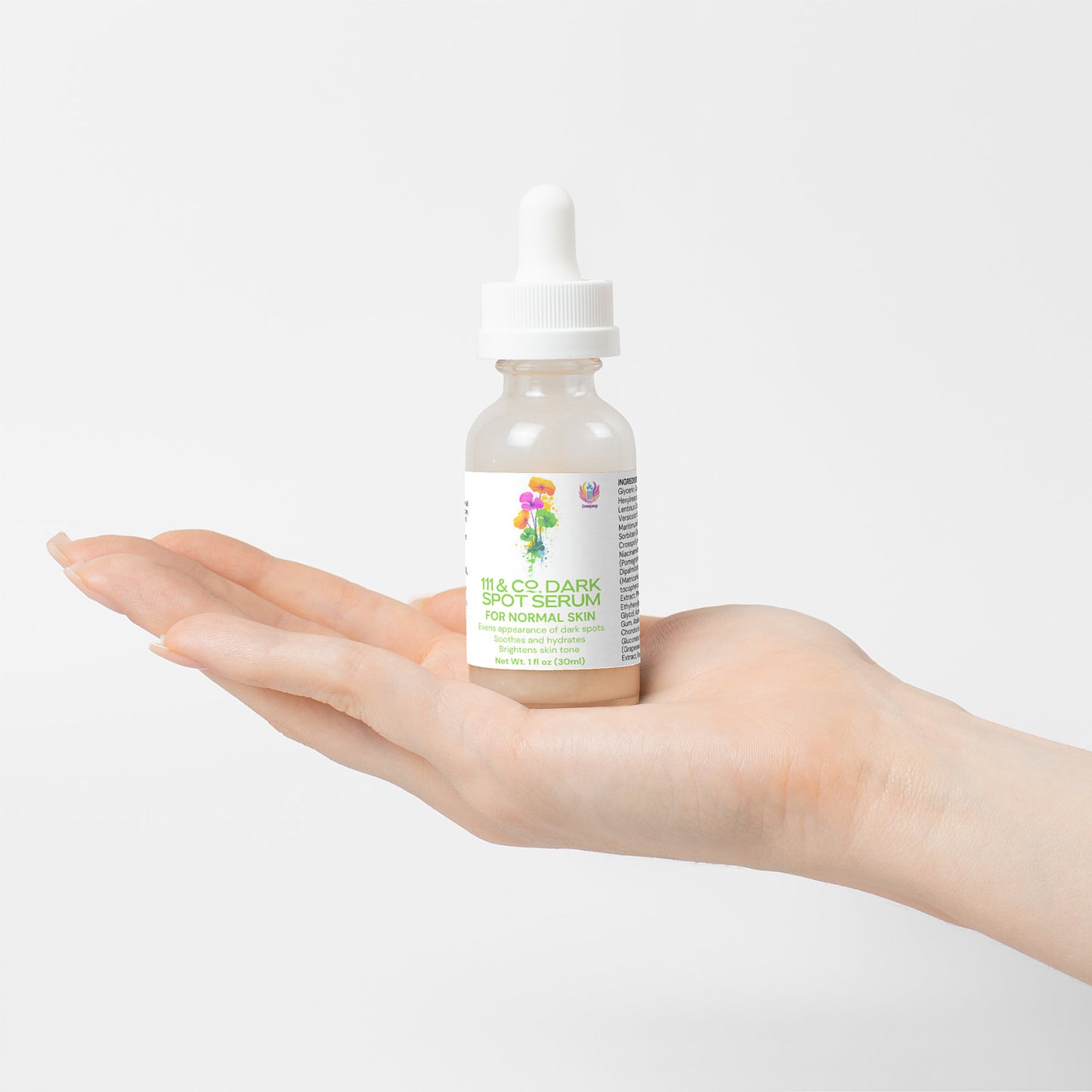 A hand holds a small bottle of 111 & Company Dark Spot Serum for Normal Skin, designed to reduce dark spots and even skin tone. The dropper cap complements the labels colorful floral graphics, making the product stand out elegantly against the plain white background.