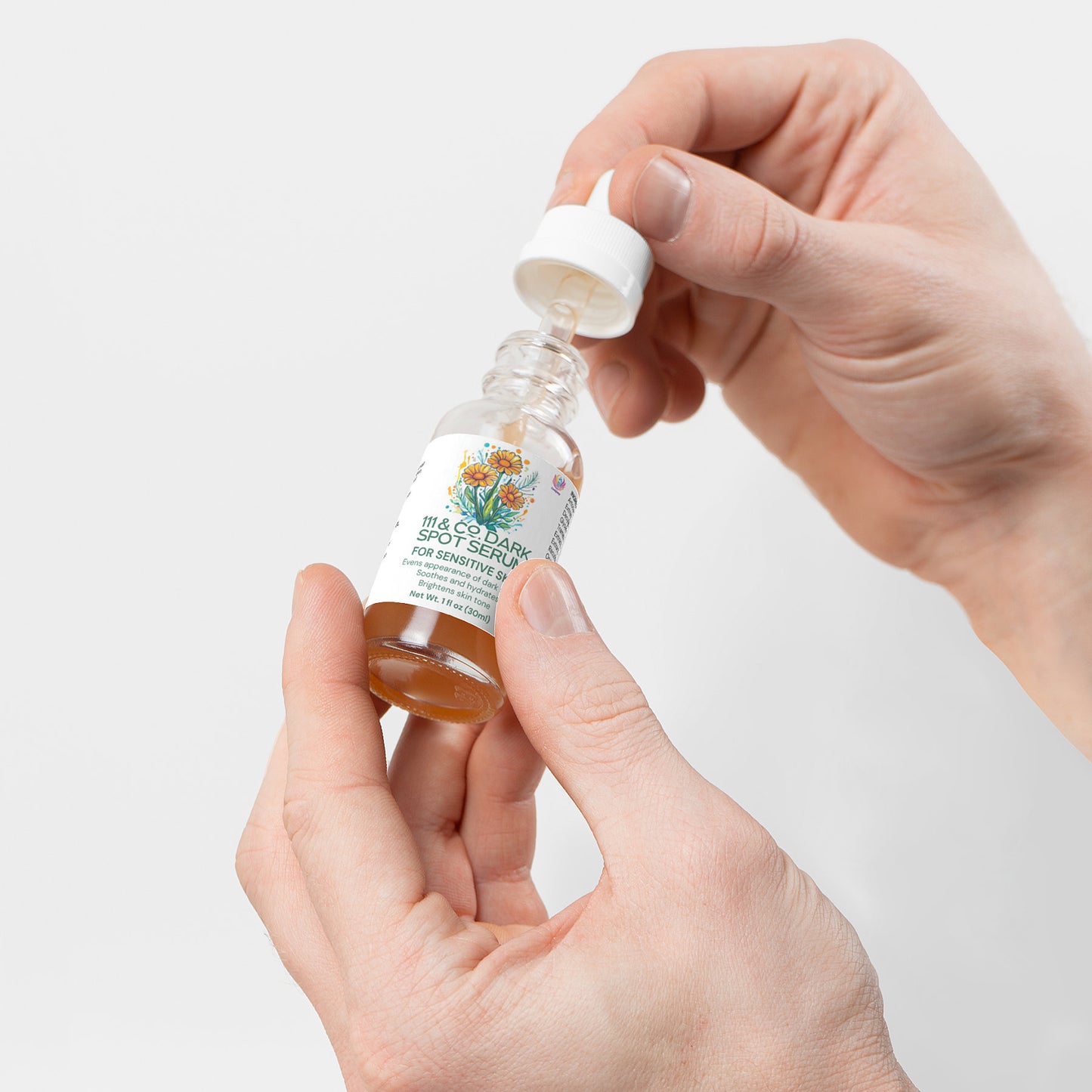 Close-up of hands holding a small bottle of face serum with a dropper. The label reads 111 & Company Dark Spot Serum for Sensitive Skin and features floral artwork. Crafted for sensitive skin, the amber-colored serum boasts natural ingredients on a plain white background.