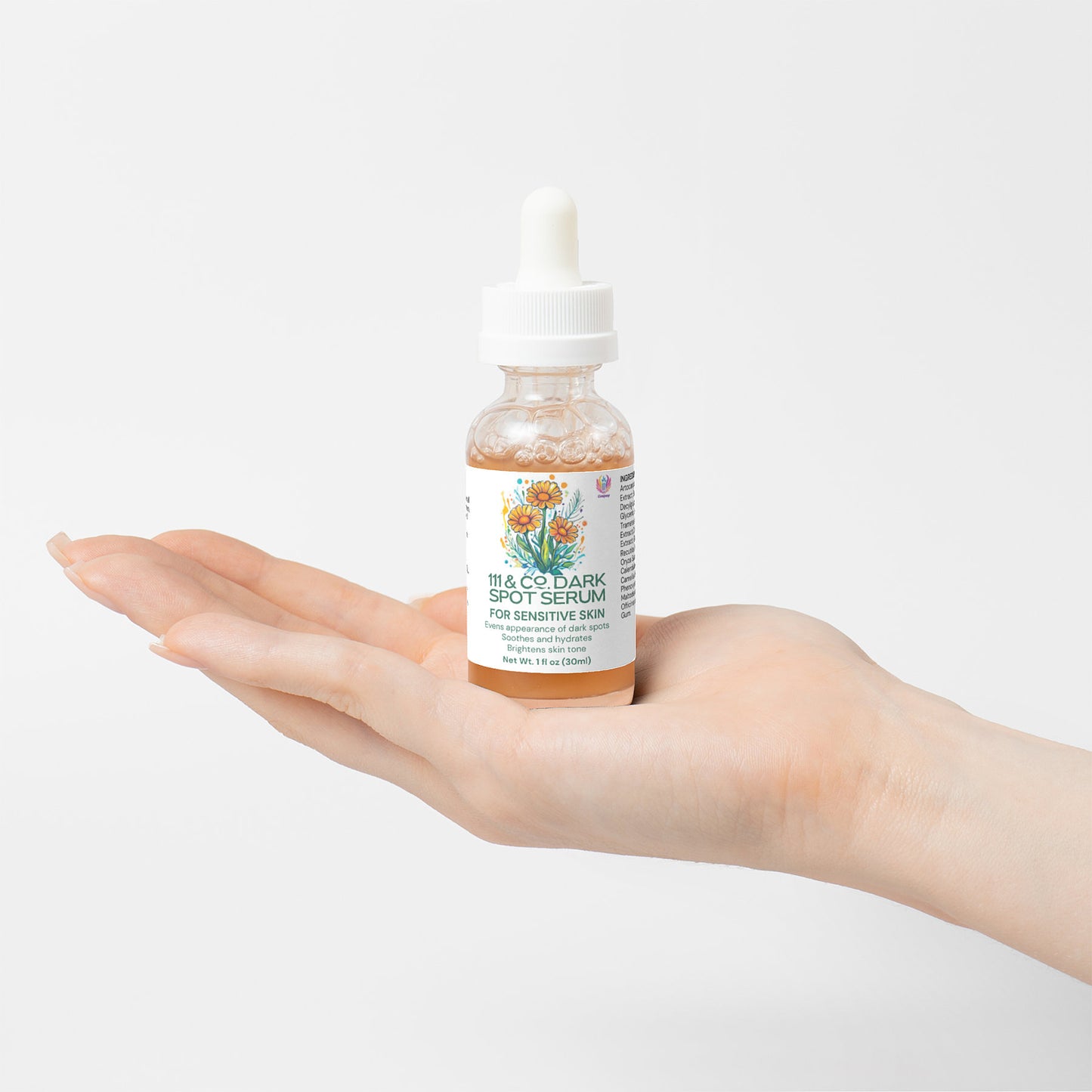 A hand holds a bottle of 111 & Company Dark Spot Serum for Sensitive Skin, crafted with natural ingredients. The glass bottle boasts a dropper and a floral illustrated label. The clear serum contrasts the plain white backdrop, emphasizing purity.