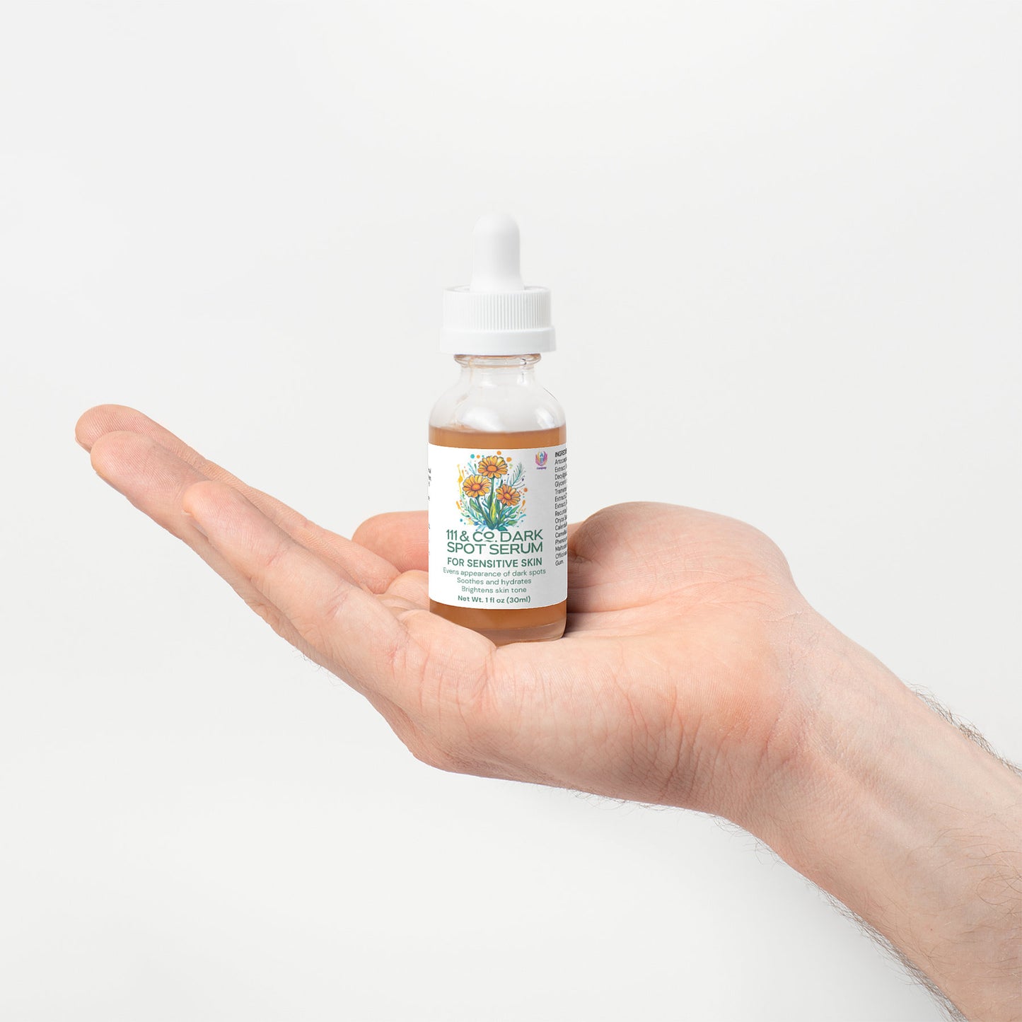A hand gracefully holds a small bottle with a dropper lid labeled 111 & COMPANY DARK SPOT SERUM FOR SENSITIVE SKIN by 111 & Company, highlighting its natural ingredients and featuring floral illustrations against a soothing, soft off-white background.