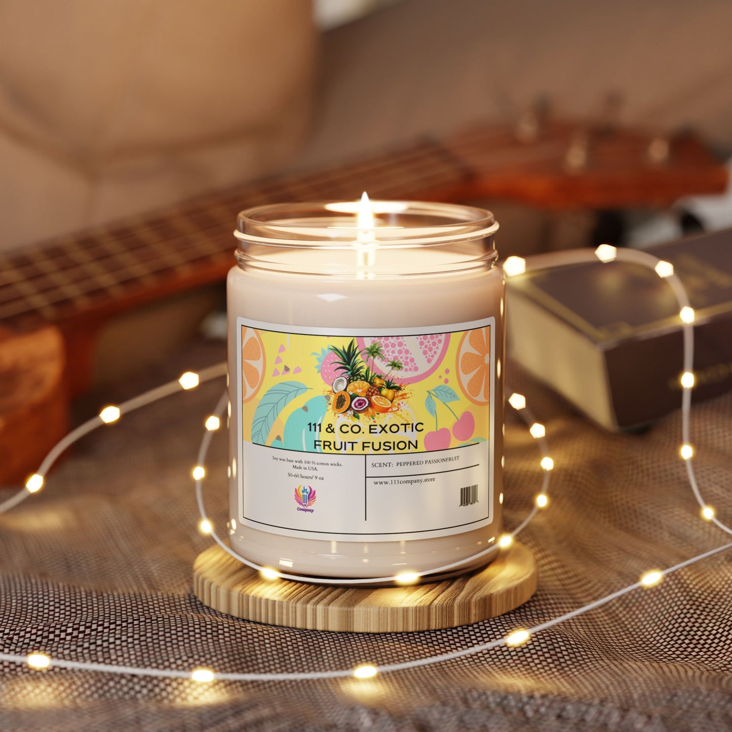 A soy wax candle in a glass jar labeled 111 & Company Exotic Fruit Fusion Peppered Passionfruit by 111 & Company offers an exotic fragrance on a wooden base. It has colorful fruit graphics, surrounded by fairy lights, a blurred guitar, and a silver box on a textured gray surface.