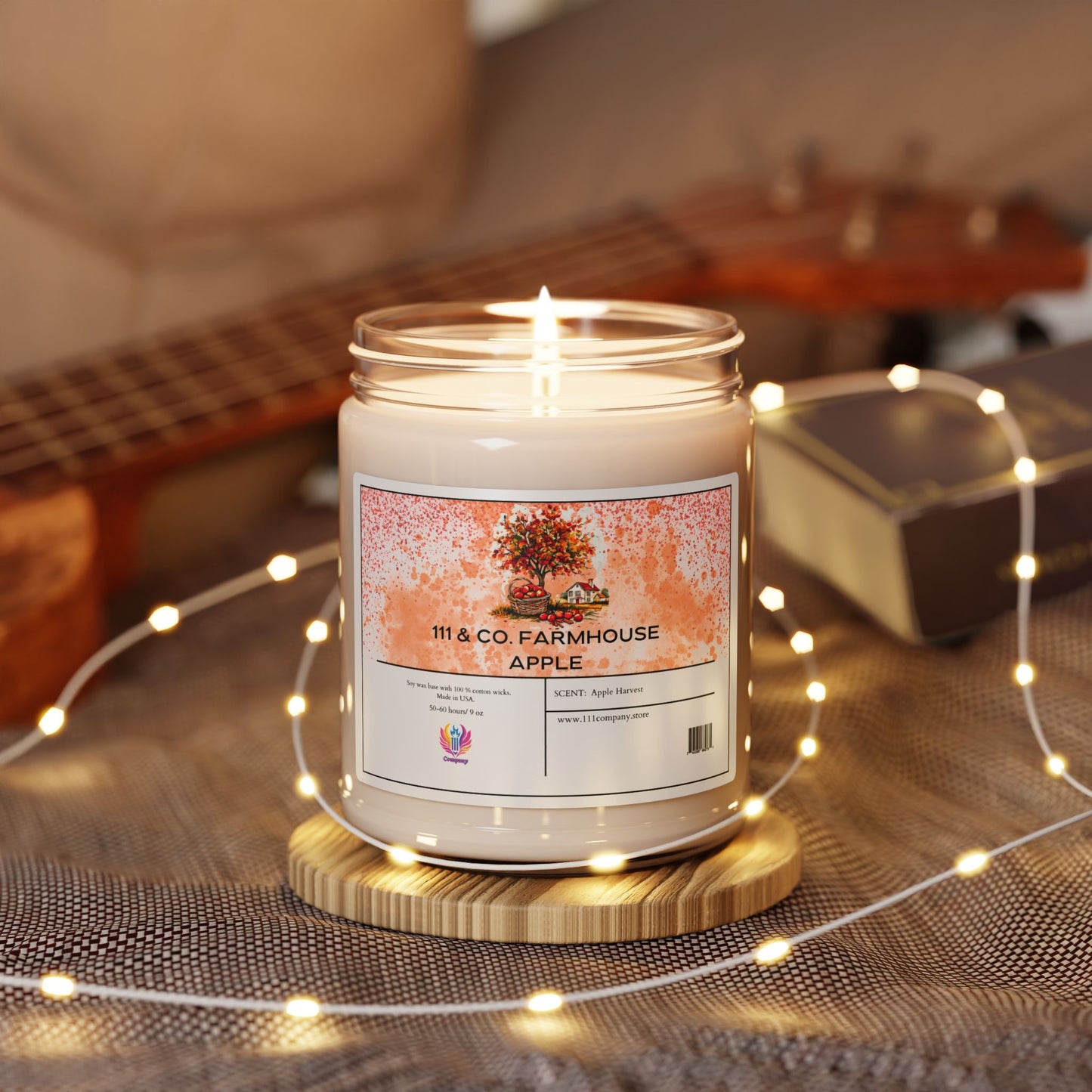A lit 111 & Company Farmhouse Apple Harvest soy candle in a glass jar rests on a wooden coaster. Surrounded by yellow fairy lights, a closed book, and a blurred ukulele, it evokes autumn with the scent of apples and flowers.