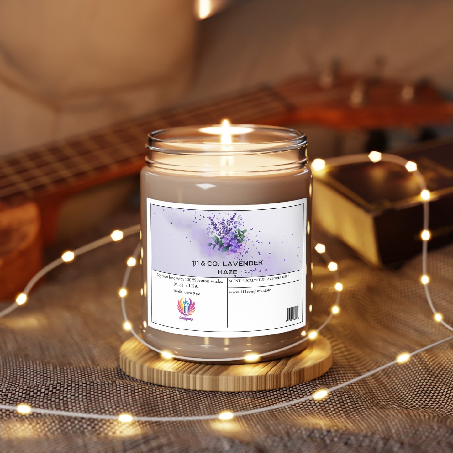 A lit 111 & Company Lavender Haze Eucalyptus Lavender Herb scented candle, soy wax 9oz by 111 & Company, rests on a wooden coaster amidst fairy lights on a textured surface. A blurred ukulele and closed book in the background add to the cozy ambiance.