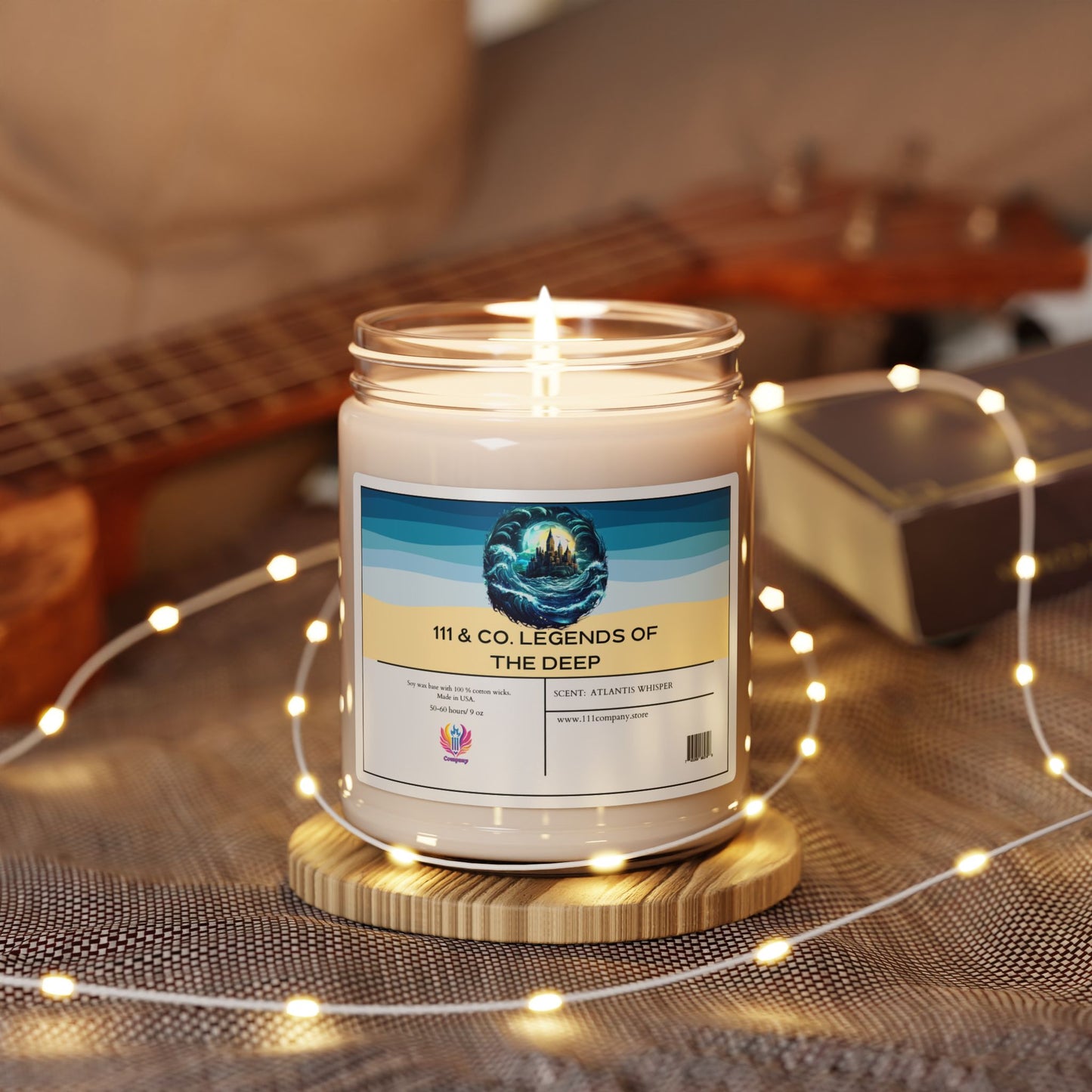 A lit 111 & COMPANY Legends of the Deep Atlantis Whisper scented candle by 111 & Company, with its ocean mist fragrance, rests on a wooden coaster surrounded by glowing fairy lights. In the background, a partially visible guitar and book sit on a gray textured surface.