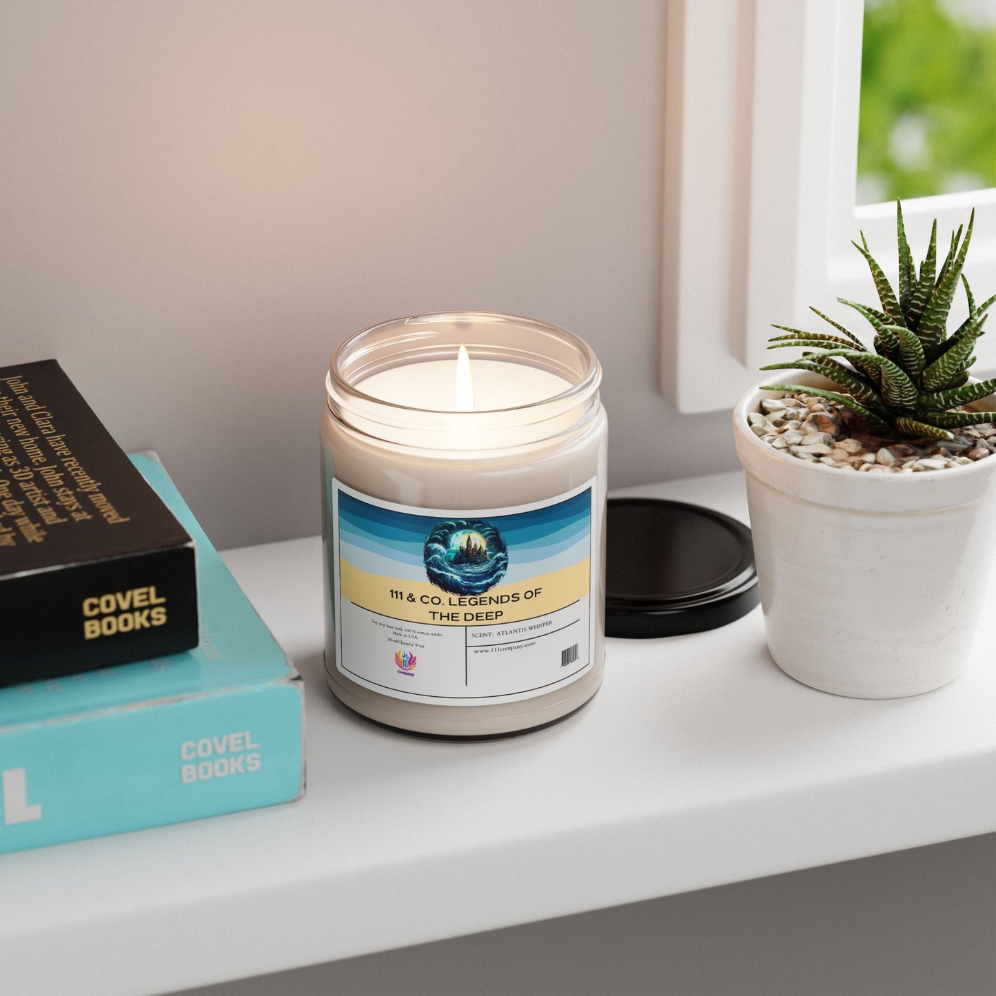 A lit 111 & Company Legends of the Deep Atlantis Whisper soy candle in a glass jar sits on a white shelf next to a potted succulent. Two teal-covered Covel Books are stacked nearby with greenery visible through the window.