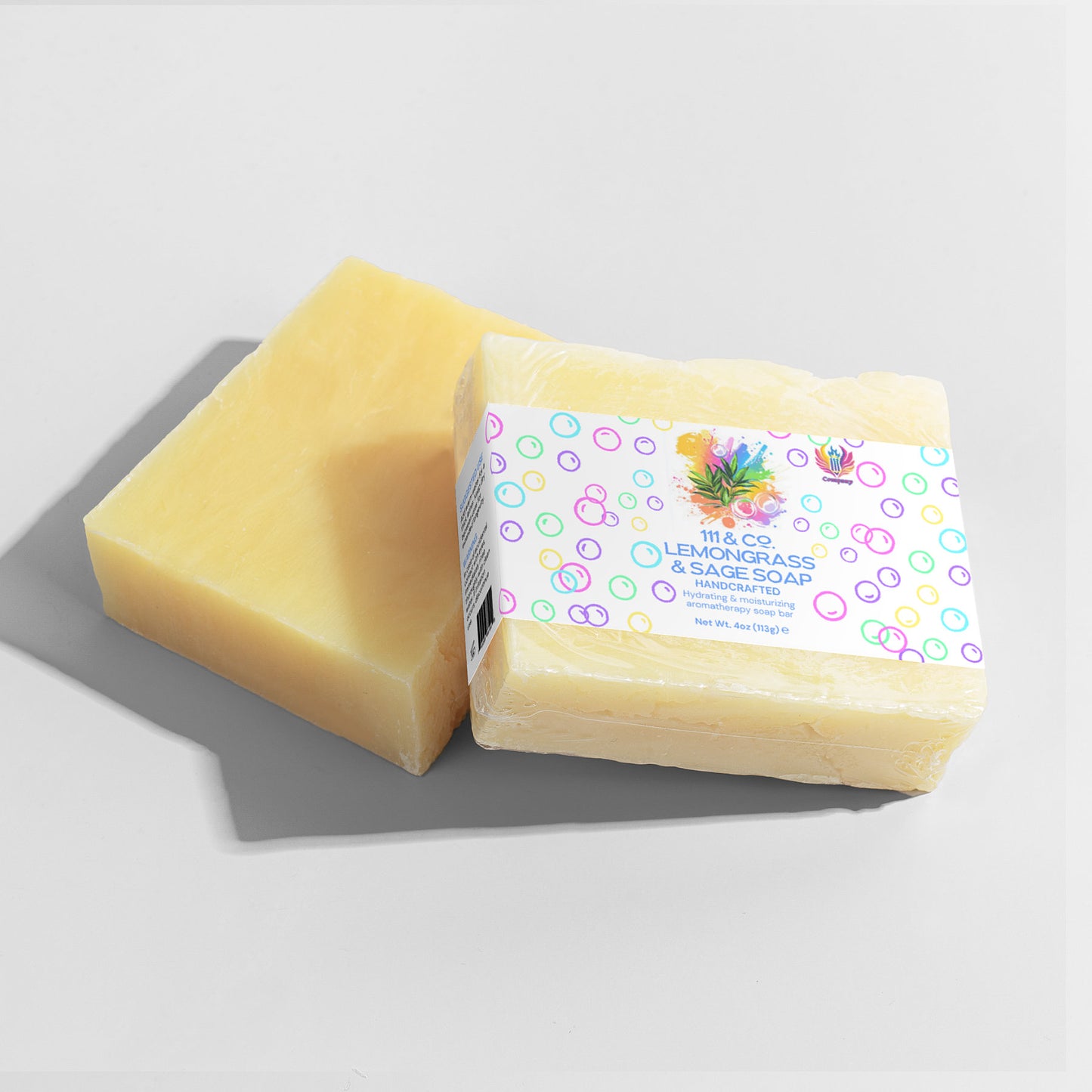 Two bars of soap lie on a light gray surface, one unwrapped with a smooth, pale yellow look ideal for sensitive skin. The other is wrapped in white paper featuring vibrant bubbles and the text: 111 & COMPANY Lemongrass & Sage Soap, Handmade. Shadows subtly cover the scene.