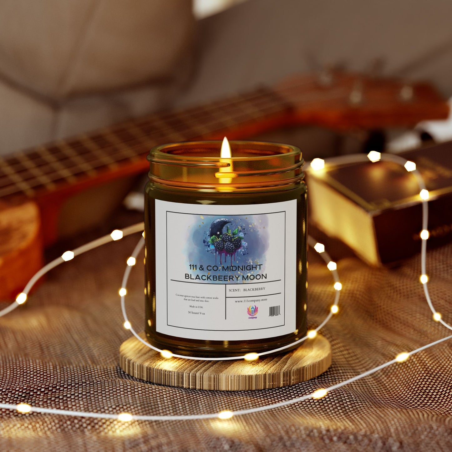 A lit 111 & Company Midnight Blackberry Moon candle in an amber jar rests on a wooden coaster. Surrounded by string lights on textured fabric, its fruity aroma fills the air. A blurred guitar and brown book in the background create a cozy, warm ambiance with eco-friendly coconut apricot wax charm.