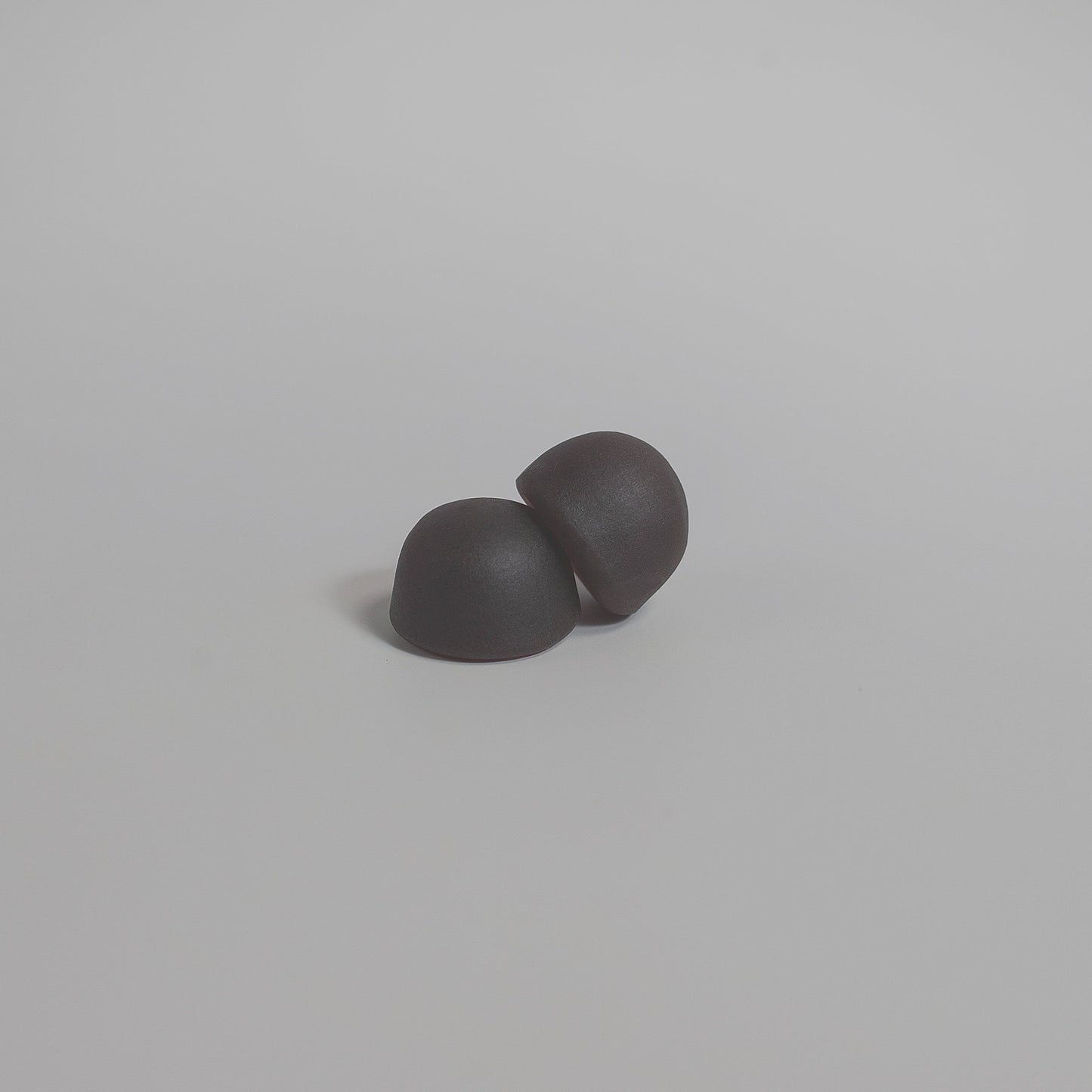 Two small, smooth, black hemispherical objects rest on a light gray surface. Like 111 & COMPANY MUSHROOM COMPLEX GUMMIES, they are positioned together, casting subtle shadows that highlight their shape and texture.