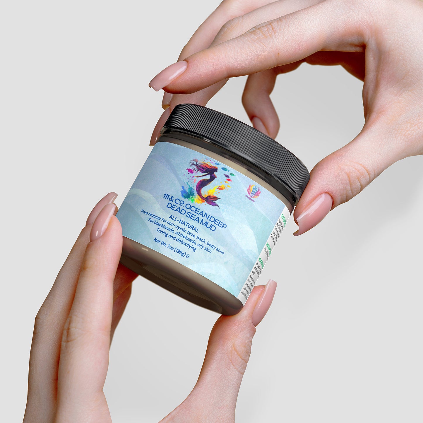 Two hands with manicured nails hold a jar of 111 & Company OCEAN DEEP DEAD SEA MUD mask from 111 & Company, highlighting its mineral-rich, 100% natural skincare formula. The label boasts a colorful abstract design, and the jar has a black lid set against a soft gray background.