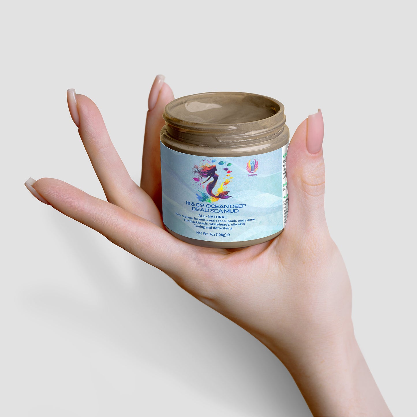 A manicured hand holds a jar of 111 & Company OCEAN DEEP DEAD SEA MUD with a colorful seahorse. The mineral-rich brown mask is visible through the open lid, set against a plain light gray backdrop, emphasizing this natural skincare gem.