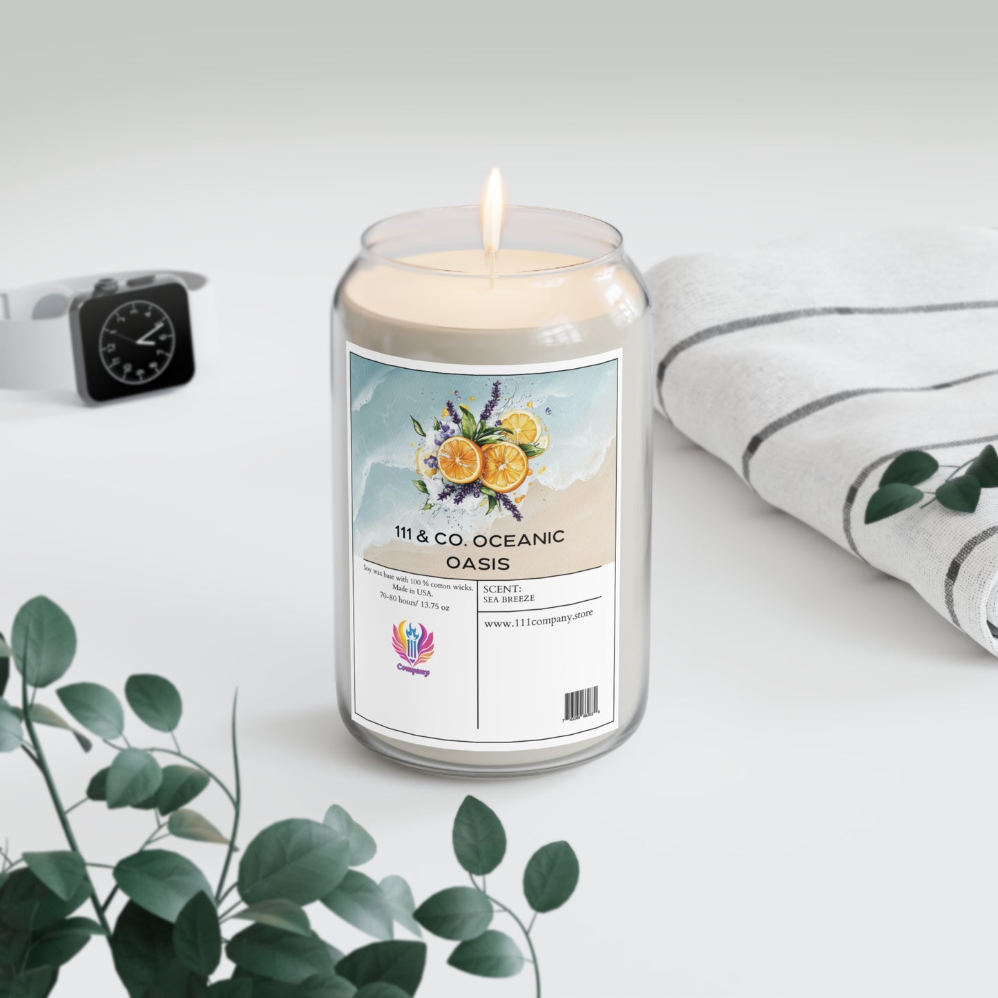 A 111 & Company Oceanic Oasis Sea Breeze soy wax candle from 111 & Company, with citrus, lavender, and cedarwood notes, sits in a glass jar on a light surface. Nearby are green eucalyptus leaves, an Apple Watch, and a striped white cloth, creating a calm and fresh ambiance.