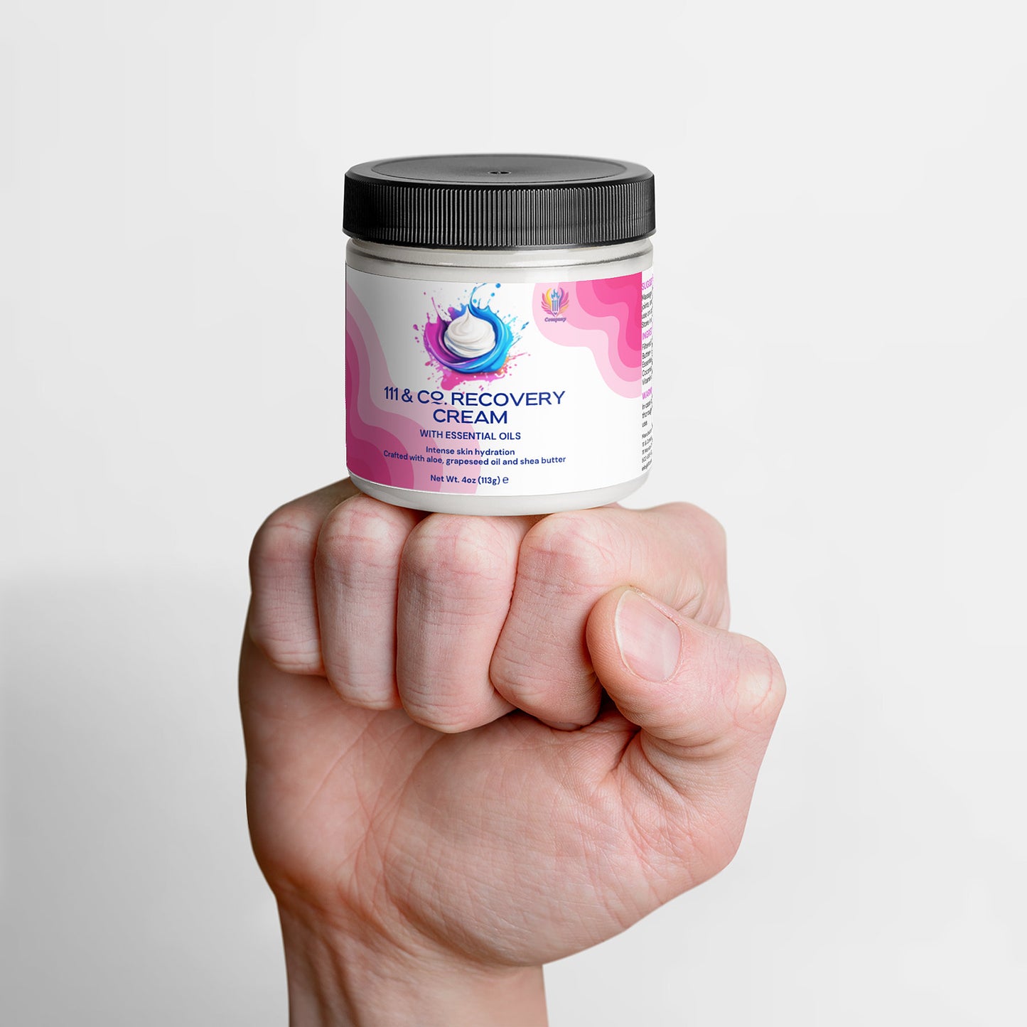 A hand holds a jar of 111 & Company Recovery Cream by 111 & Company, infused with essential oils, ashwagandha, and shea butter for superior skin hydration. The white and pink label features a stylized water splash design and is topped with a black lid against a white background.