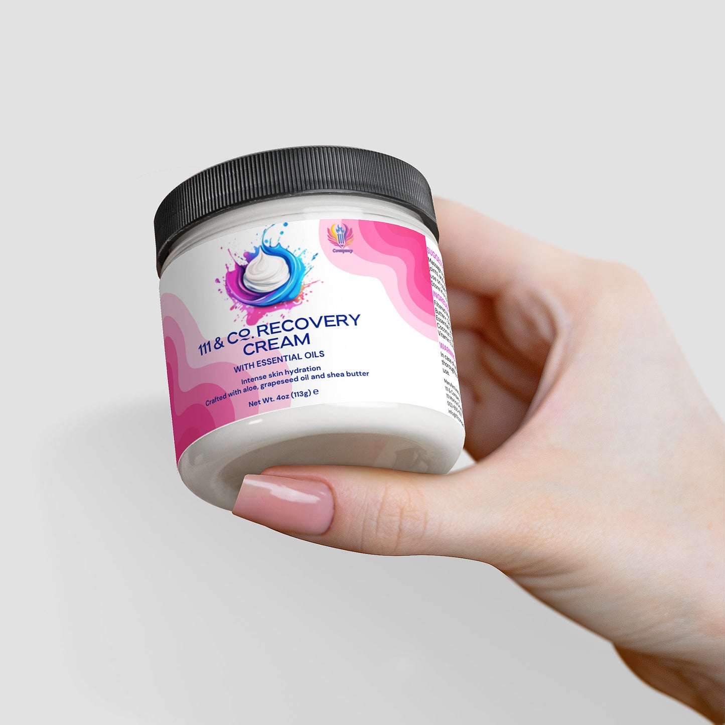 A light-skinned hand holds a jar of 111 & COMPANY RECOVERY CREAM by 111 & Company. The white jar has a pink and blue label, highlighting ingredients like essential oils, shea butter, and aloe for hydration. A plain light gray background accentuates the product.