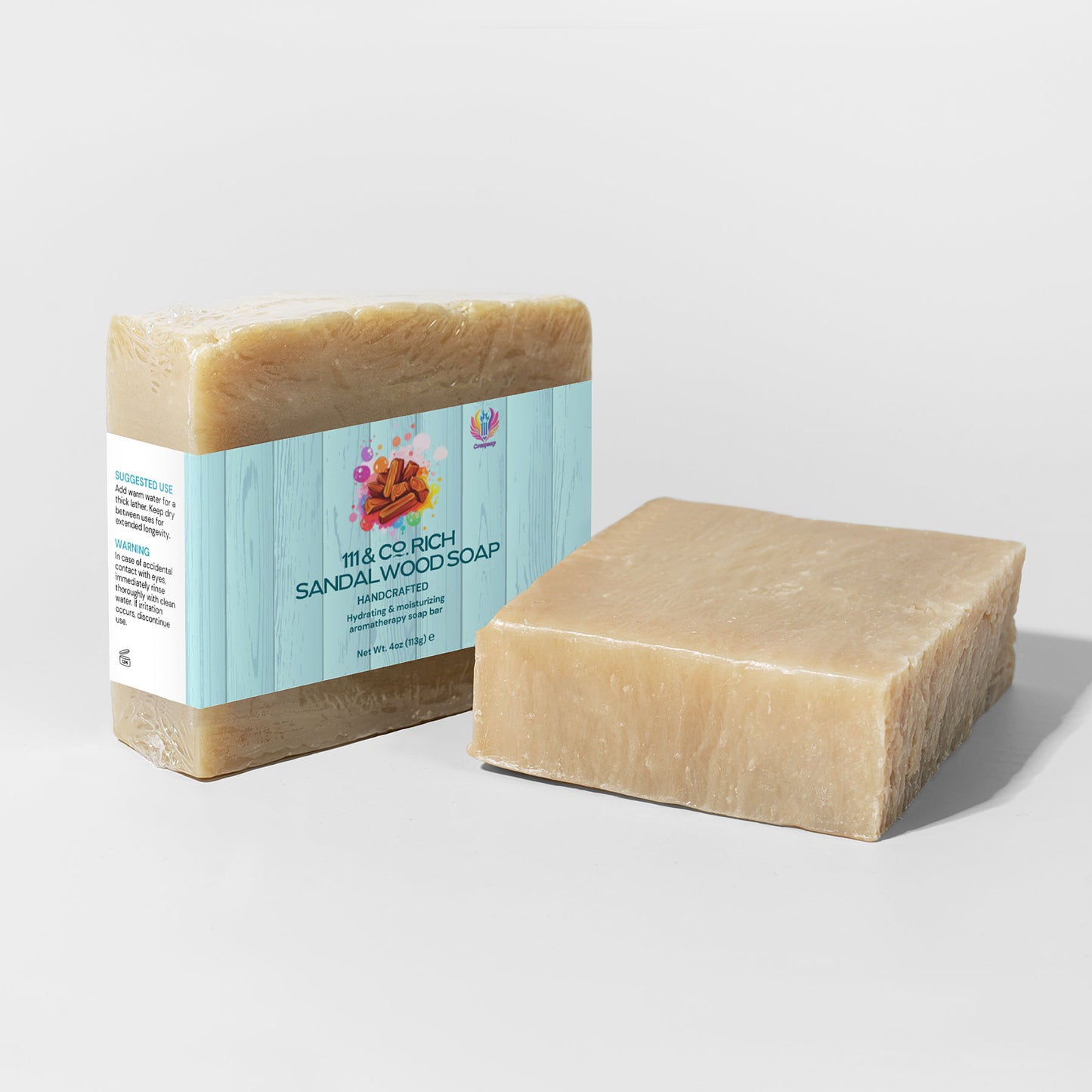 Two tan soap bars; one wrapped, one unwrapped. The wrapped bar has a light blue label with a floral design reading 111 & COMPANY RICH SANDALWOOD SOAP, Handcrafted by 111 & Company. Ideal for sensitive skin, both are rectangular with smooth edges on a pale gray surface.