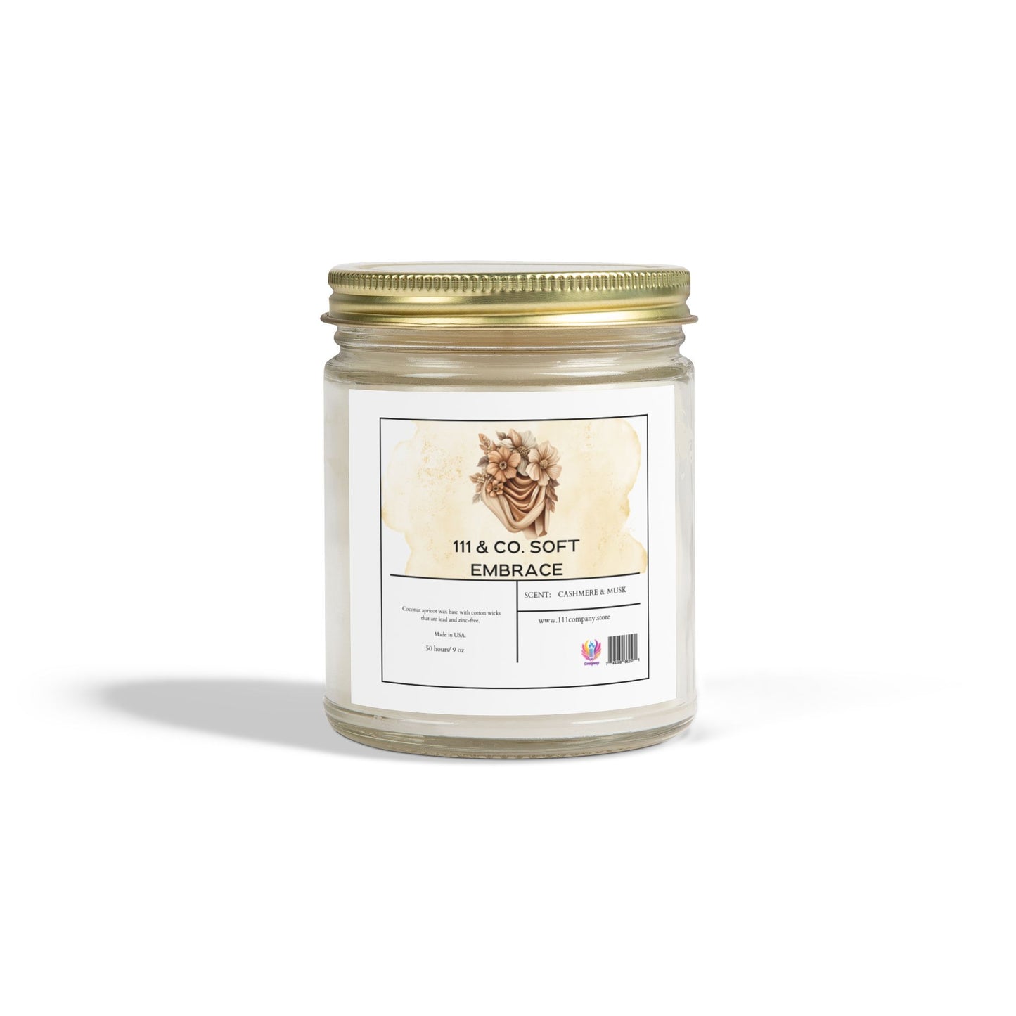 The 111 & Co. Soft Embrace candle in a clear jar offers a cashmere musk scent with vanilla notes. Made from eco-friendly coconut apricot wax, it boasts a gold metal lid, floral design label, and provides 50 hours of burn time against a plain white background.