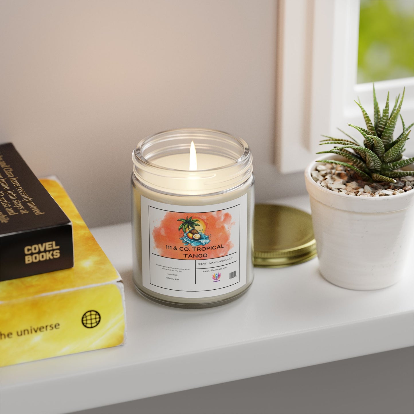 A lit 111 & Company candle labeled 111 & Company Tropical Tango emits a coconut-mango scent on a white surface beside a succulent and books. Made from eco-friendly coconut apricot wax, the candle casts a yellow glow with its green lid nearby, as natural light streams through a window.