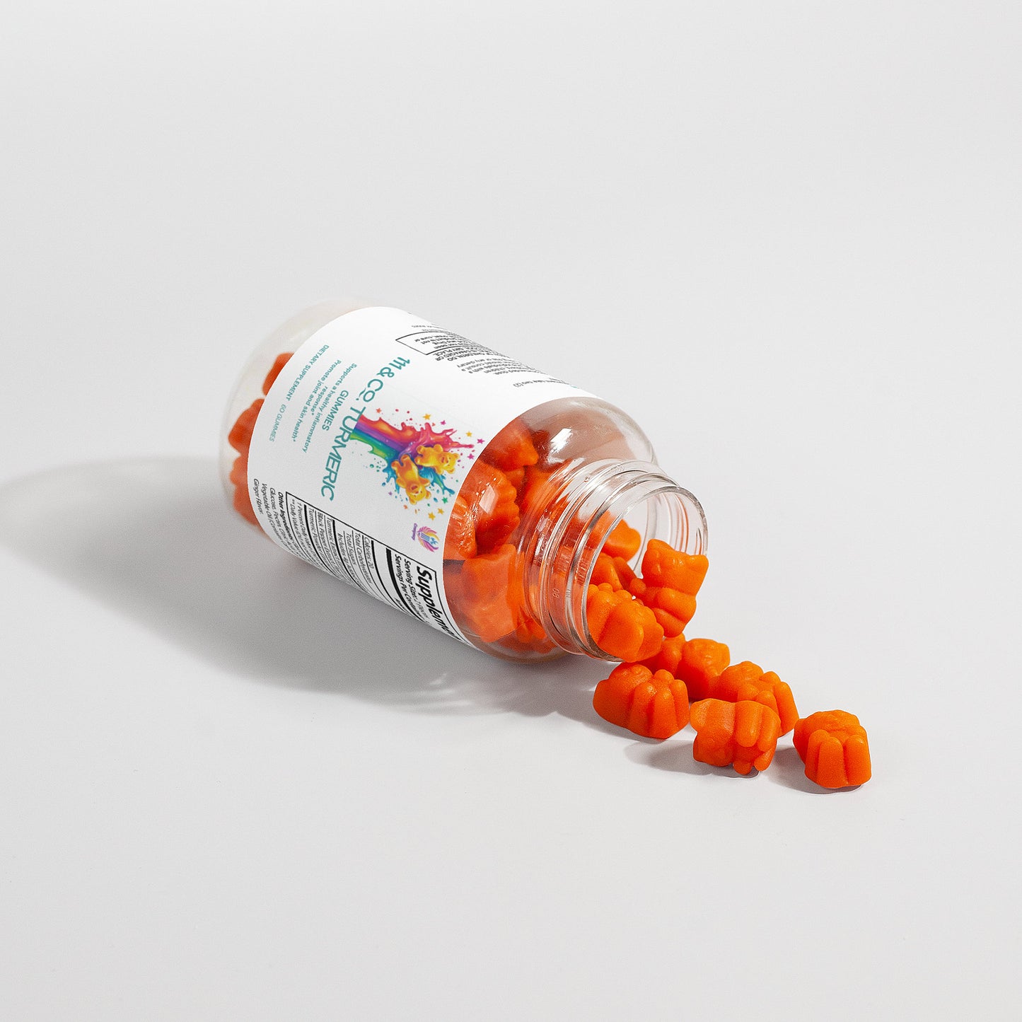A clear plastic bottle of 111 & COMPANY TURMERIC GUMMIES lies on its side with the lid off, spilling pumpkin-shaped gummies. The colorful label highlights their antioxidant benefits as shadows cast to the left enhance the vibrant contents on a plain white surface.
