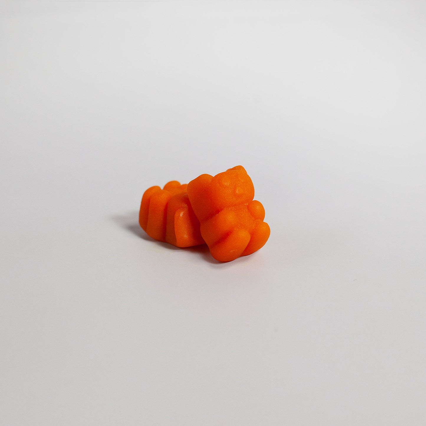 Two 111 & COMPANY TURMERIC GUMMIES by 111 & Company sit closely on a smooth white surface. The lighting reveals their translucent texture and curcumin hint, casting a soft shadow and highlighting the vibrant color and playful shape, exuding a minimalist vibe.