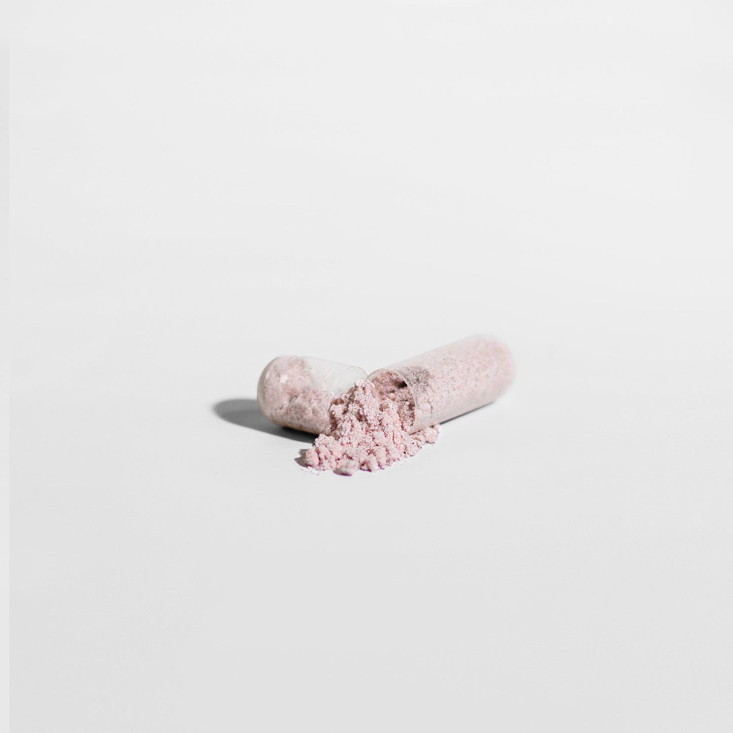 A close-up of an 111 & COMPANY VISION SUPPORT CAPSULE by 111 & Company on a white background, showing light pink powder spilling out. The cylindrical capsule, split into two halves, lies horizontally with the powder forming a small pile and casting a soft shadow.