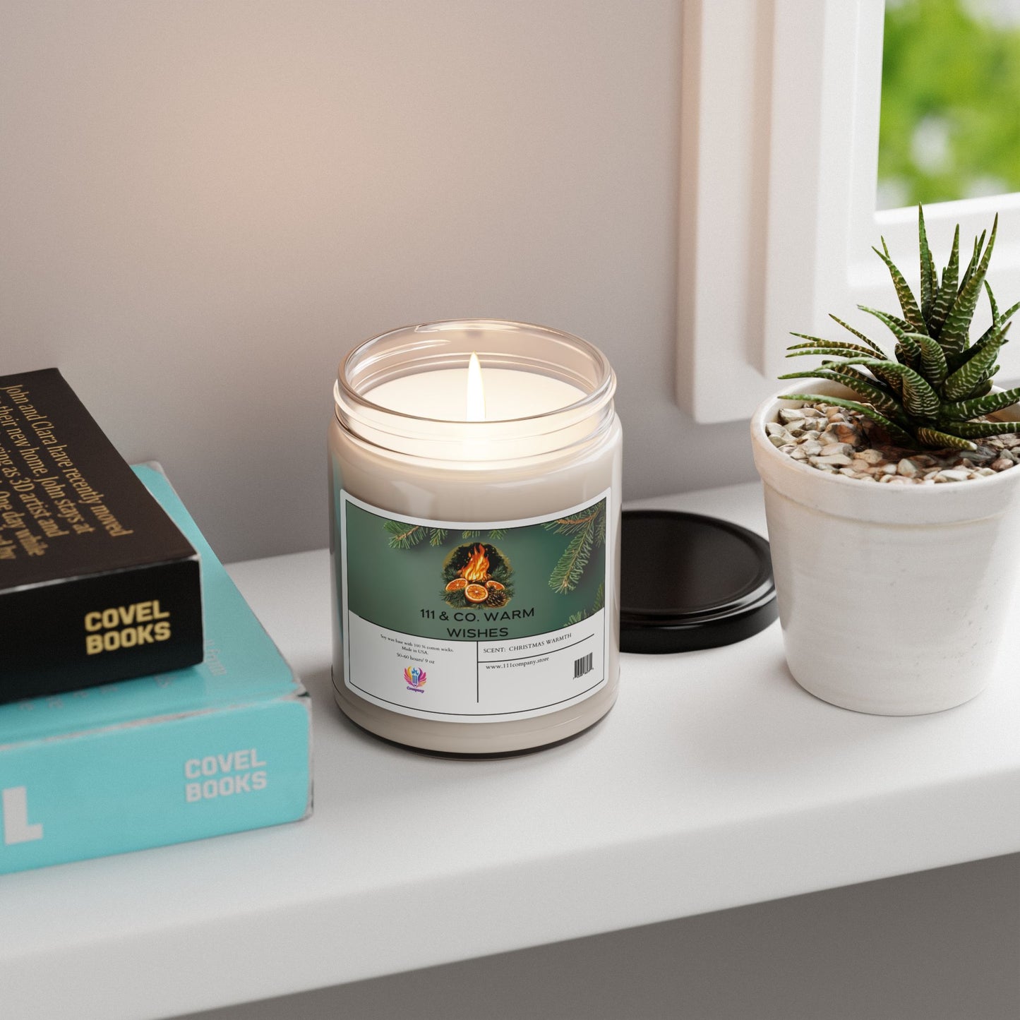A lit white 9oz 111 & Company soy wax candle brings Christmas warmth to a white shelf, nestled beside a potted succulent and stacked COVEL BOOKS. The label, featuring pine cones and greenery, reads 111 & COMPANY WARM WISHES, with its black lid nearby, capturing the holiday fragrance essence.