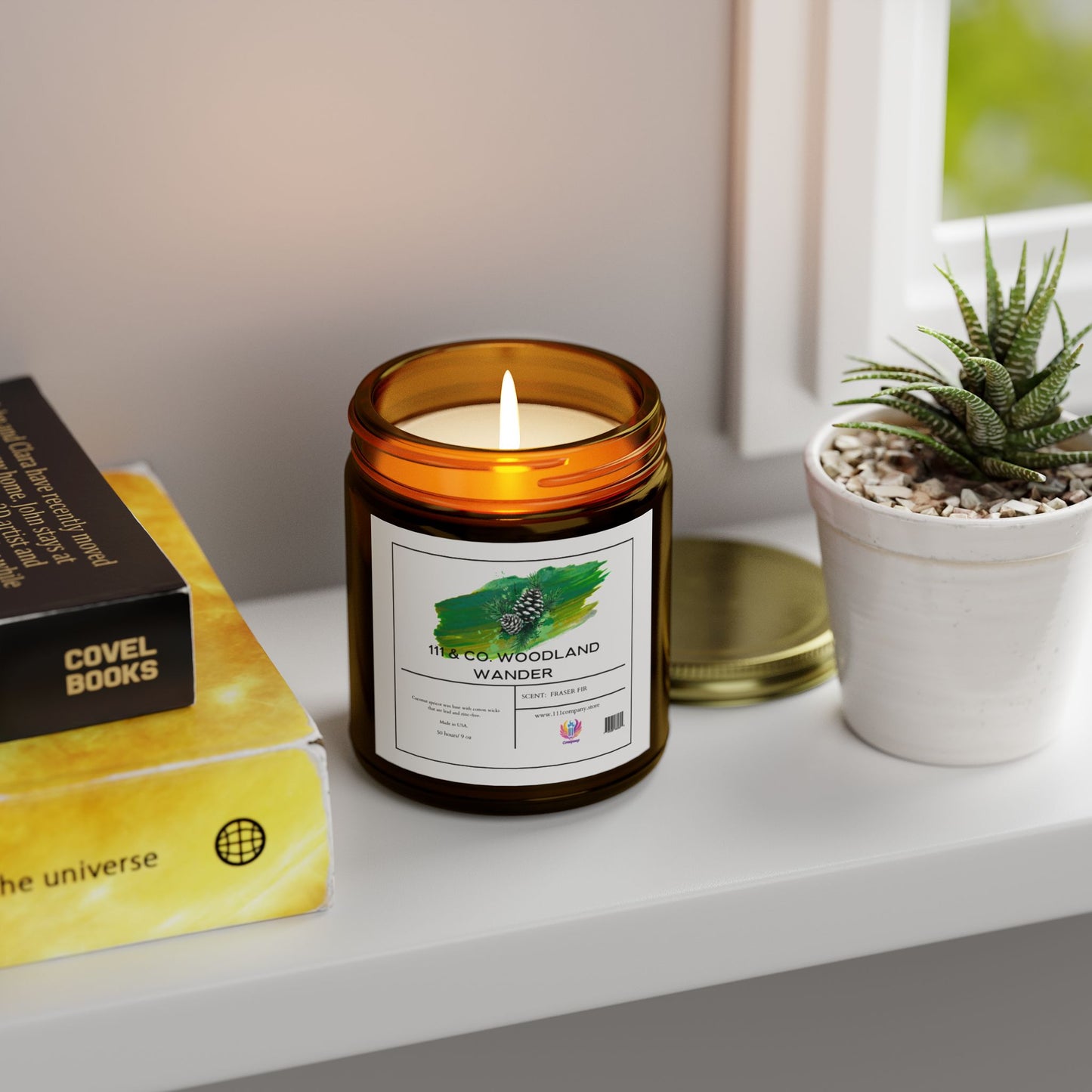 A lit 111 & COMPANY Woodland Wander Fraser Fir candle from 111 & Company, made of Coconut Apricot Wax and housed in a brown jar, sits on a white shelf beside a potted succulent. Nearby are two books—one black, one yellow—under soft natural light from a closed window.
