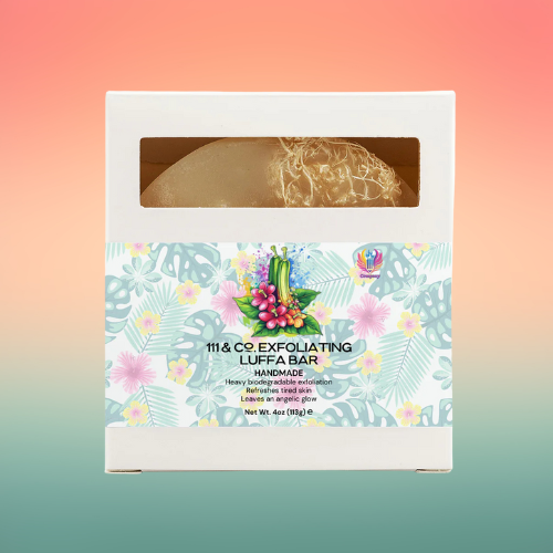 Packed in a white box with tropical leaf, flower, and fruit designs, the 111 & Company Exfoliating Luffa Bar is a handmade skincare bar with visible loofah inside—ideal for natural skin care fans. Background features a soft pink and green gradient.