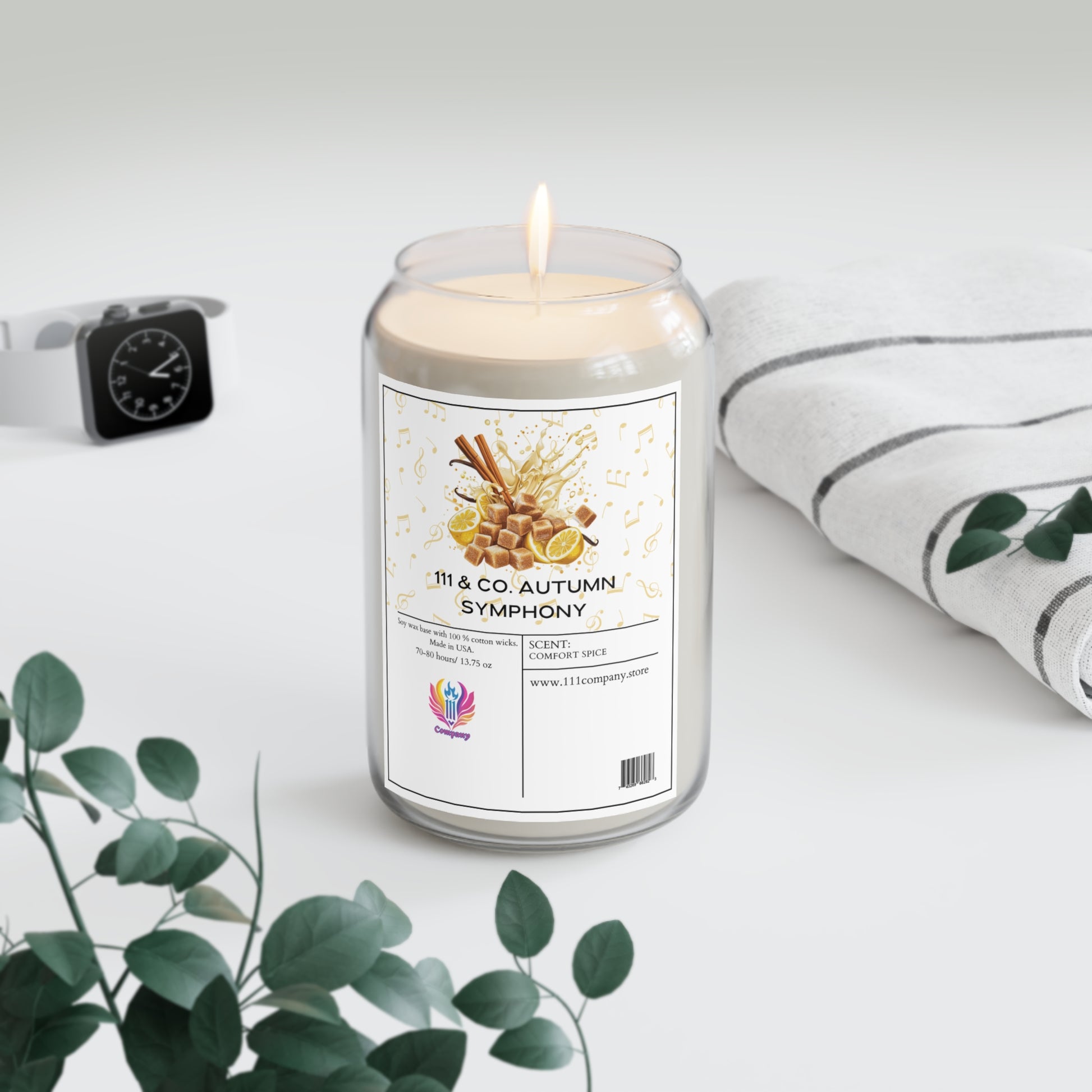 A lit 111 & COMPANY AUTUMN SYMPHONY Comfort Spice candle in a glass jar by Printify creates a cozy ambiance. Its label, featuring honeycomb and cinnamon sticks, is eco-friendly. Nearby, a folded striped cloth, smartwatch, and eucalyptus sprigs complement the light gray background.