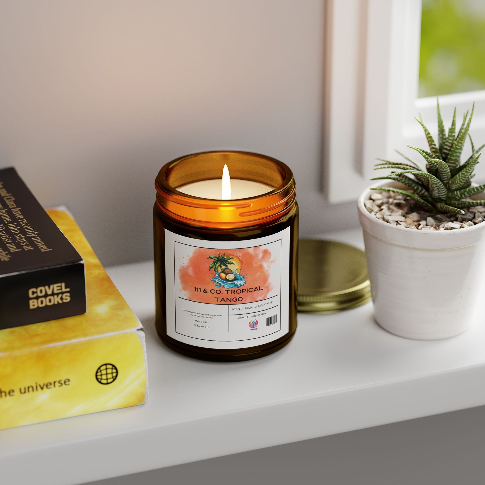 A 111 & COMPANY Tropical Tango coconut mango candle from Printify infuses the air with fragrance as it sits lit in its amber jar on a white shelf. Nearby, a succulent and The Universe book stack complement the scene, with its gold lid beside the eco-friendly 9oz coconut apricot wax candle.