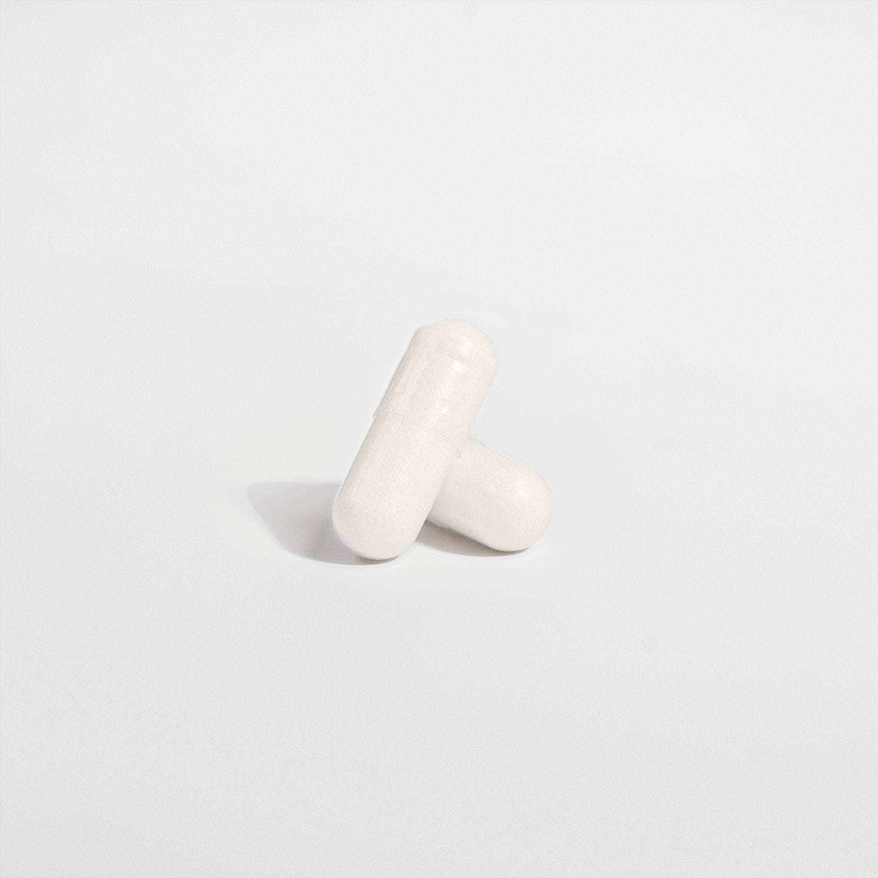 Two white capsules of 111 & Co. 5-HTP by 111 & Company form a cross on a plain white background, casting soft shadows for a clean, minimalist look, promoting serotonin support and enhancing emotional well-being.