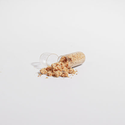 The 111 & COMPANY BEE PEARL capsule is open on a white surface, spilling its beige vitamin-rich powder. Finely textured granules slightly scatter around it, forming a small mound. Subtle shadows accentuate the minimalistic scene.
