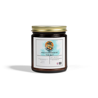 The 111 & Company Breeze by the Bay amber jar candle, crafted with coconut apricot wax and featuring a beachwood scent, includes a white-labeled seashell and palm tree design on a blue watercolor background with a gold lid.