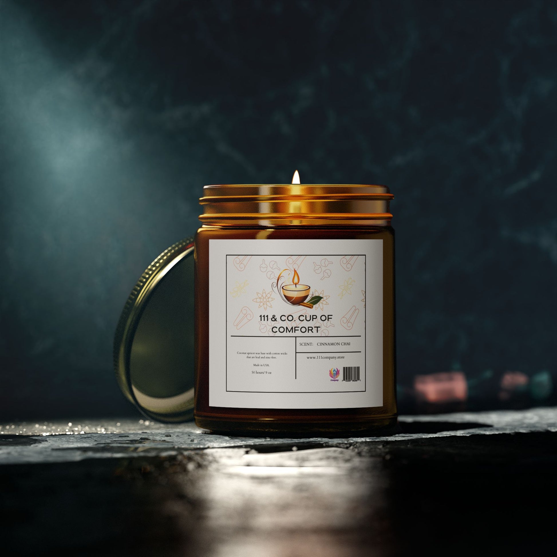 An amber glass candle labeled 111 & COMPANY CUP OF COMFORT CINNAMON CHAI, made with natural Coconut Apricot Wax, sits on a dark surface. Featuring a steaming cup graphic, this 9oz candle by 111 & Company emits a soft glow. The brass lid rests open behind the reusable jar.