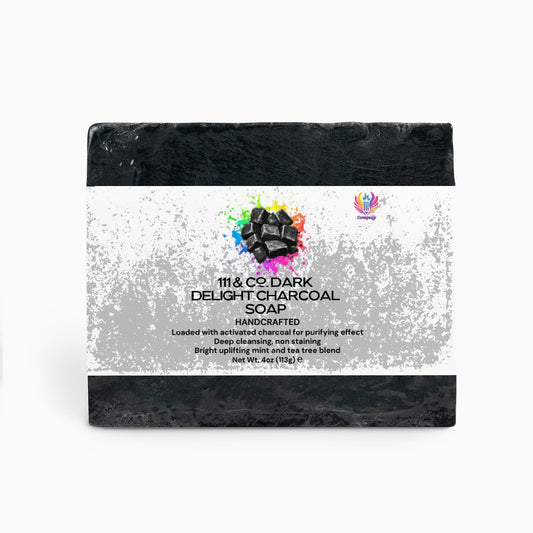 The 111 & Company Dark Delight Charcoal Soap is a rectangular bar with a white paper label featuring watercolor splashes and an image of activated charcoal. It highlights its deep cleansing, purifying effect, is handcrafted, non-staining, and includes a soothing tea tree blend.