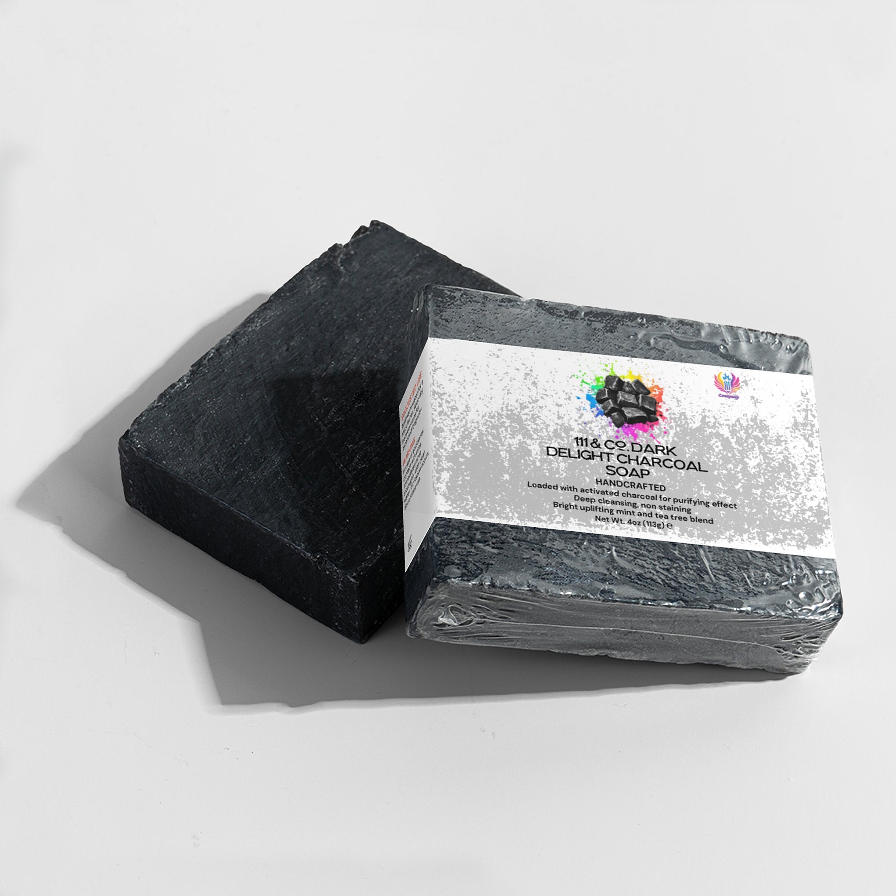 Two rectangular bars of 111 & COMPANY DARK DELIGHT CHARCOAL SOAP are shown. One is unwrapped, displaying its matte, textured surface, while the other is wrapped in a white label with colorful accents detailing the activated charcoals ingredients and features against a light gray background.