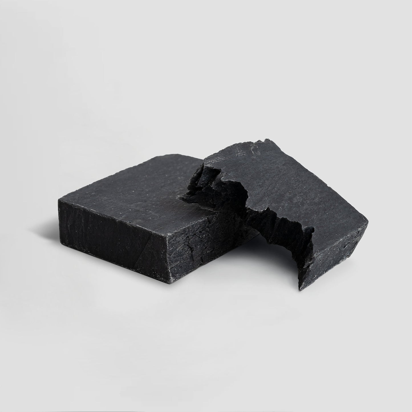 Two rectangular black bars of 111 & COMPANY DARK DELIGHT CHARCOAL SOAP, one broken into two pieces, rest on a light gray background. The rough, matte texture and uneven edges are visible in the soft lighting, highlighting subtle details.