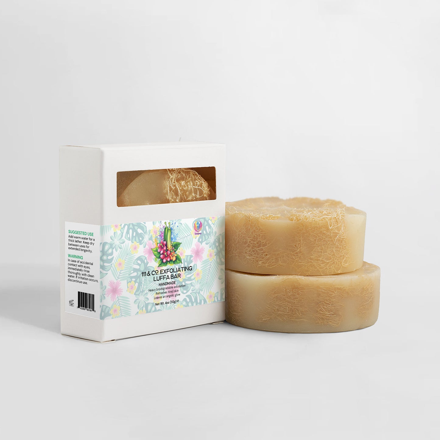 Two round luffa soaps sit beside a white box with a transparent window showing a third soap. The box has a floral design and the text 111 & Company Exfoliating Luffa Bar. Ideal for eco-friendly skin care, this biodegradable set is set against a light gray background.