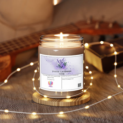 A lit 111 & Company Lavender Haze Eucalyptus Lavender Herb scented candle, soy wax 9oz by 111 & Company, rests on a wooden coaster amidst fairy lights on a textured surface. A blurred ukulele and closed book in the background add to the cozy ambiance.