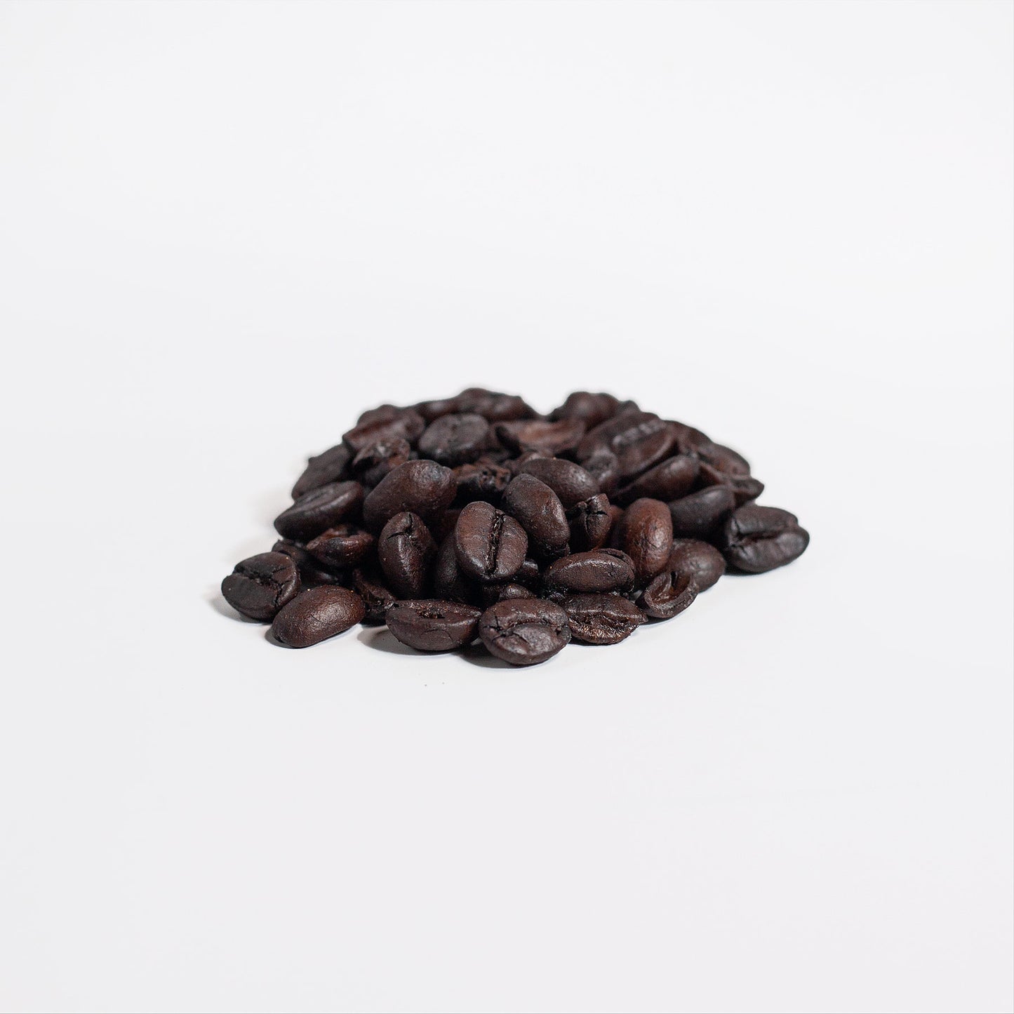 Dark roasted Arabica beans from 111 & Companys Manuka Honey Coffee 16oz are scattered on white, their shiny, oily texture reflecting light and highlighting a rich brown color. This scene evokes the aroma and depth of the coffee, with the plain background emphasizing the beans.