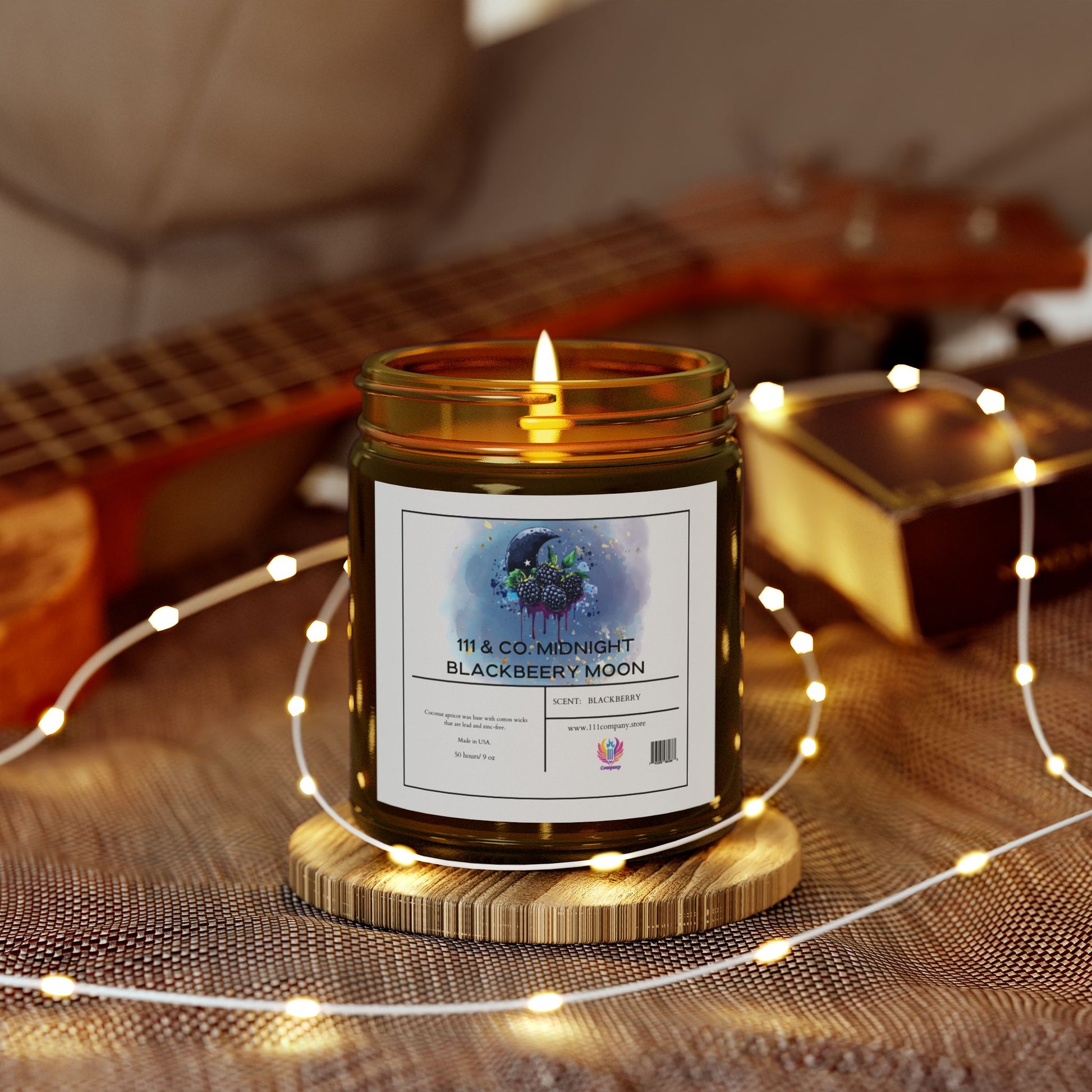 A lit 111 & Company Midnight Blackberry Moon candle in an amber jar rests on a wooden coaster. Surrounded by string lights on textured fabric, its fruity aroma fills the air. A blurred guitar and brown book in the background create a cozy, warm ambiance with eco-friendly coconut apricot wax charm.